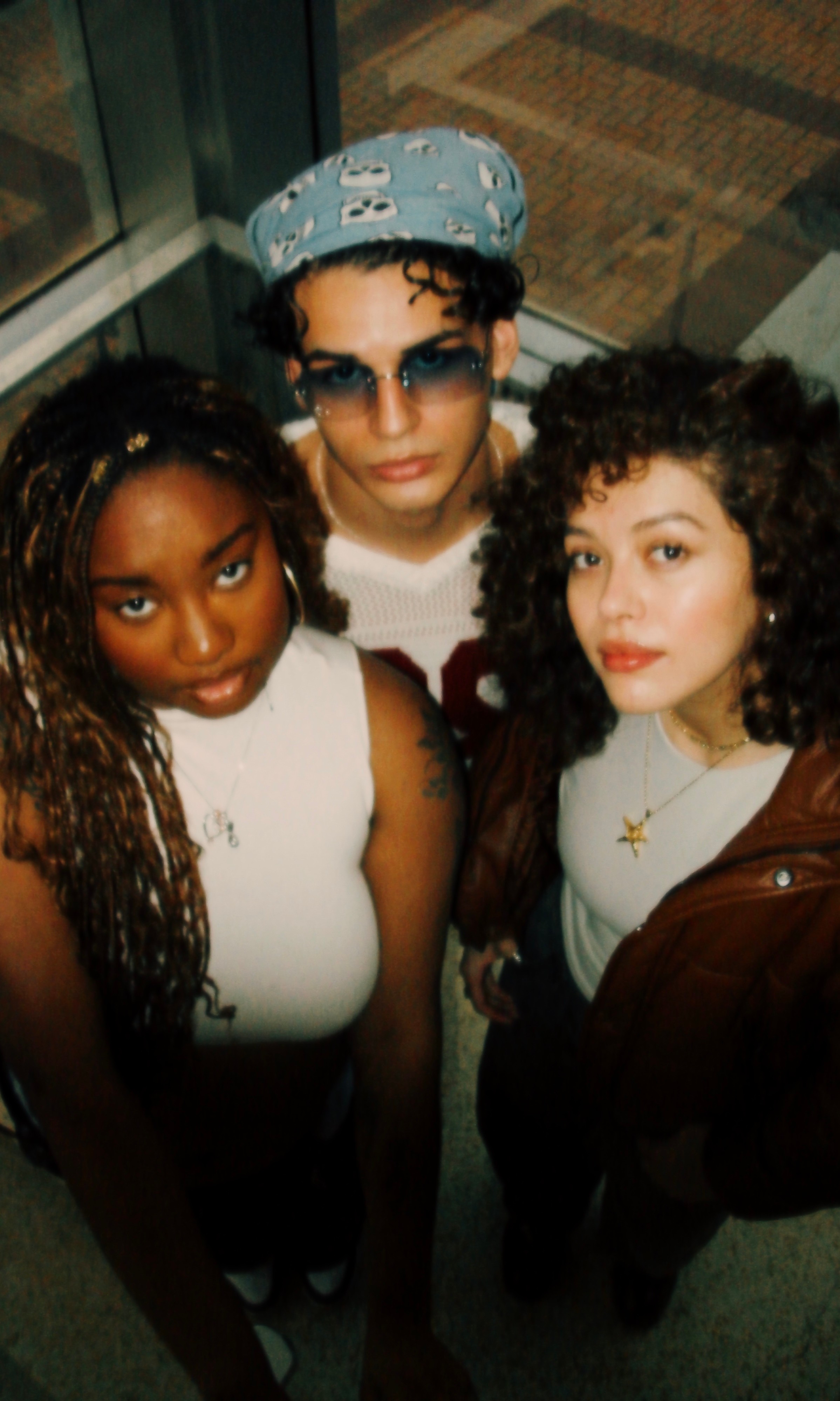 Three women standing together indoors, one wearing sunglasses and a headscarf, others with curly hair, all looking at the camera.