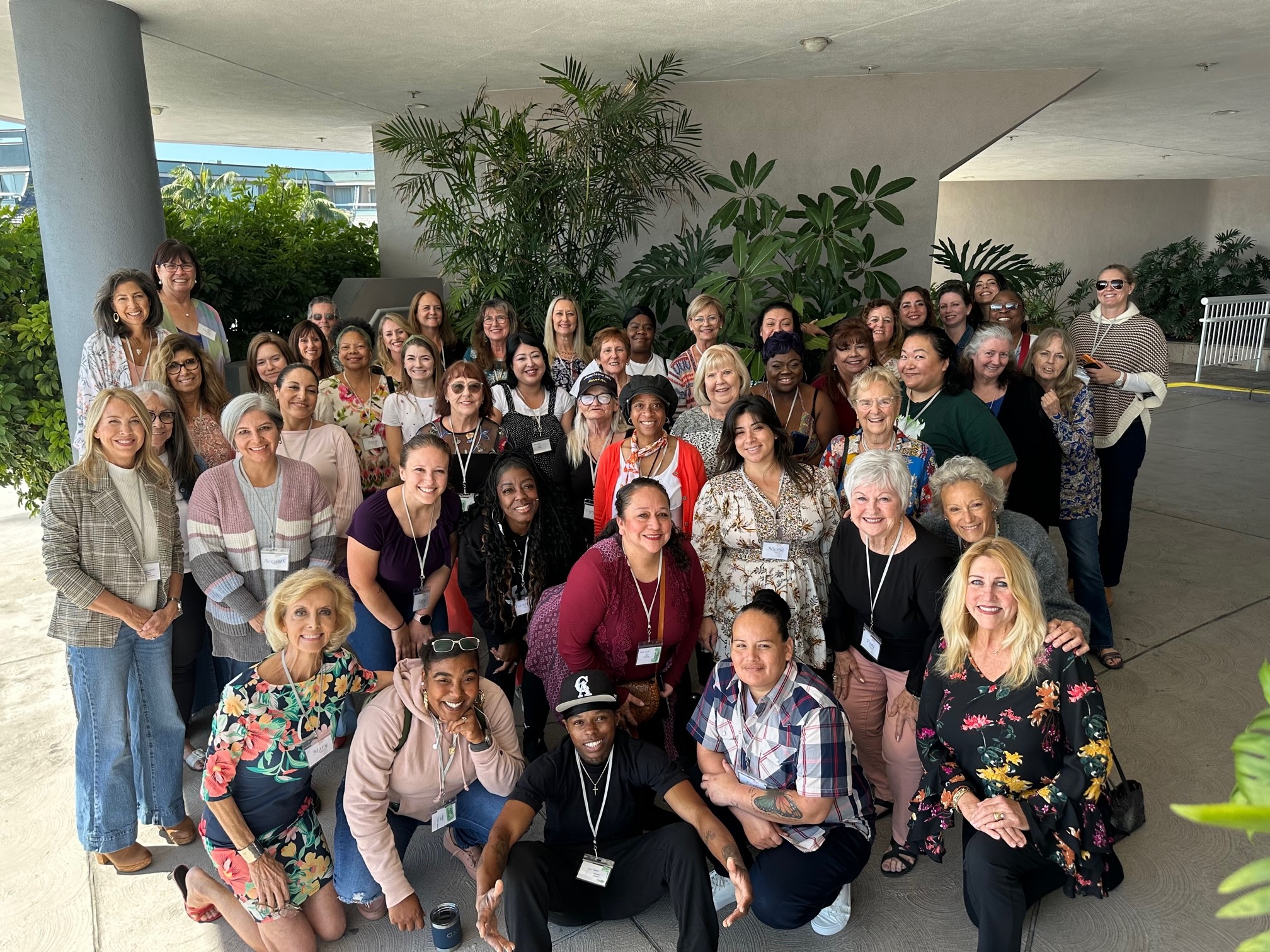 Group of diverse women and men posing outdoors under a covered area with plants in the background.