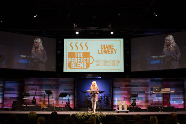 A woman stands on stage with a microphone, three screens behind her display event logos, and musical instruments are on stage.