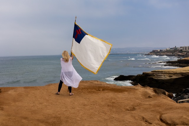 Person holding a large flag on a beach with rocks and ocean in the background, cloudy sky, wearing a white dress.