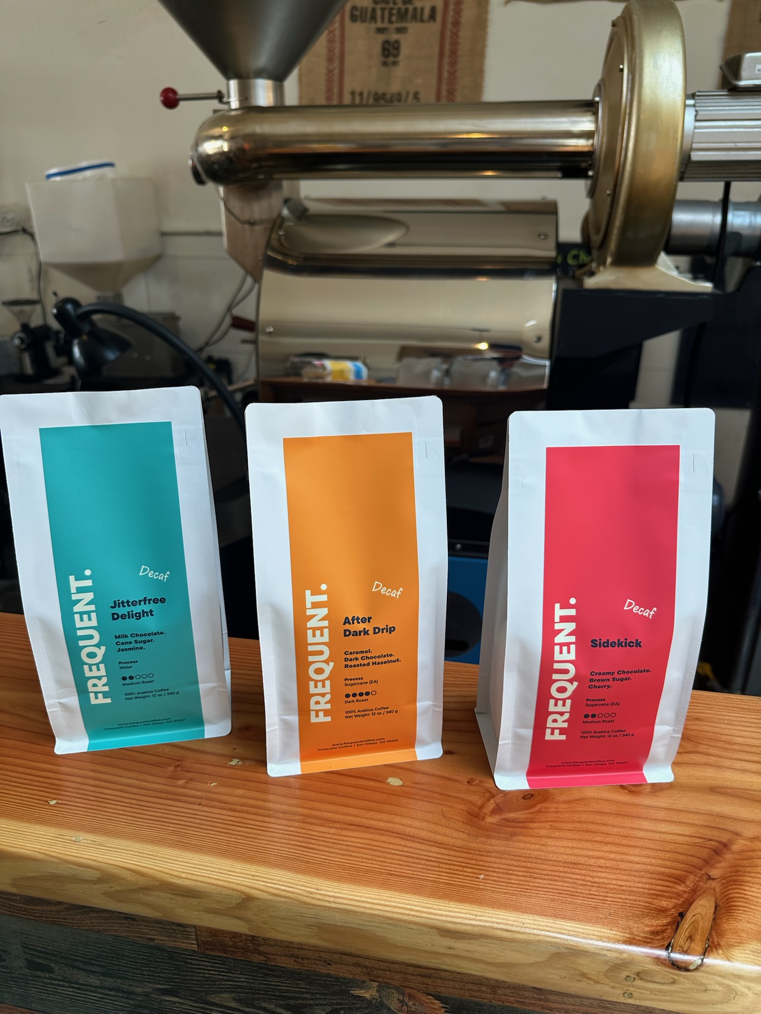 Three colorful coffee bags on a wooden surface with coffee equipment in the background.