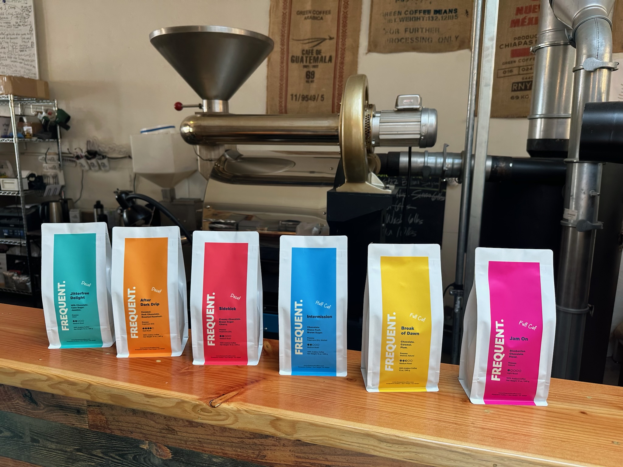 Six colorful coffee bags lined up on a wooden counter with coffee equipment in the background.