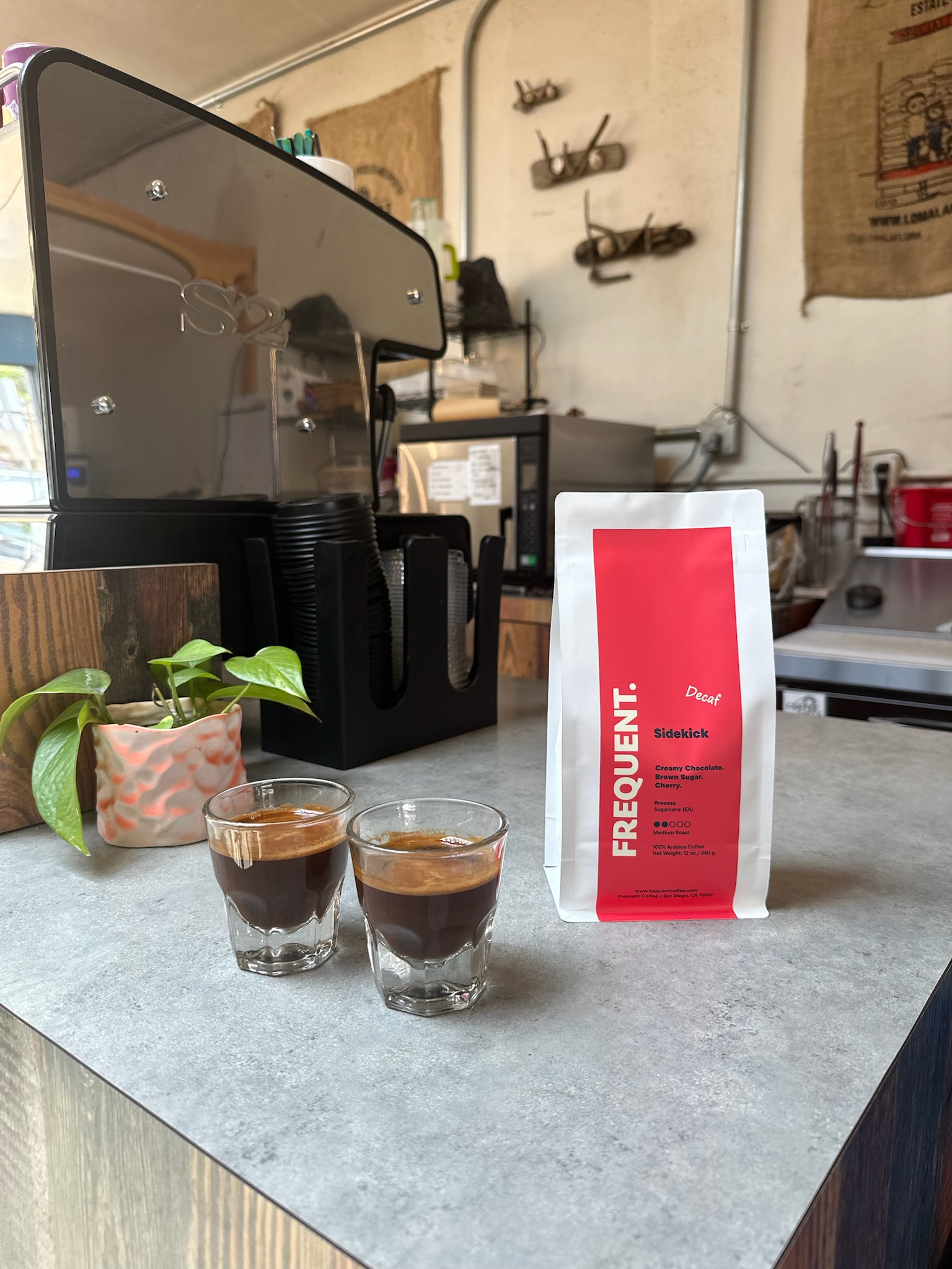Two small glasses of coffee and a red and white bag of coffee beans on a counter in a cafe, with coffee equipment and wall decorations in background.