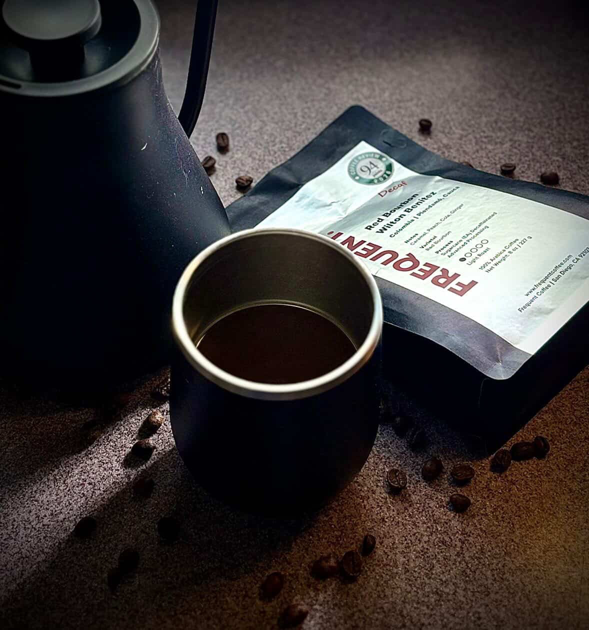 A black mug filled with coffee, a black kettle, and a packet of coffee on a brown surface with scattered coffee beans.