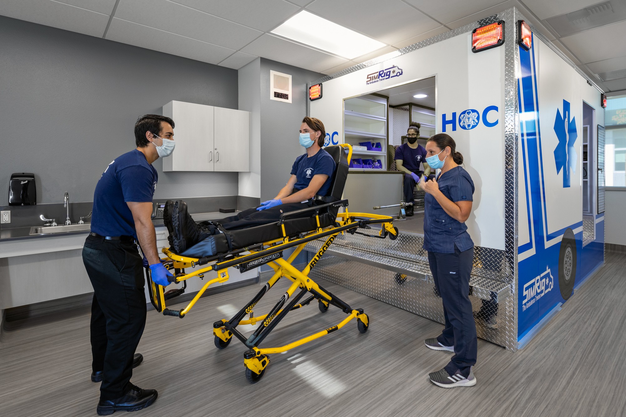 Four medical professionals in masks and scrubs prepare a patient on a stretcher outside an emergency room entrance.