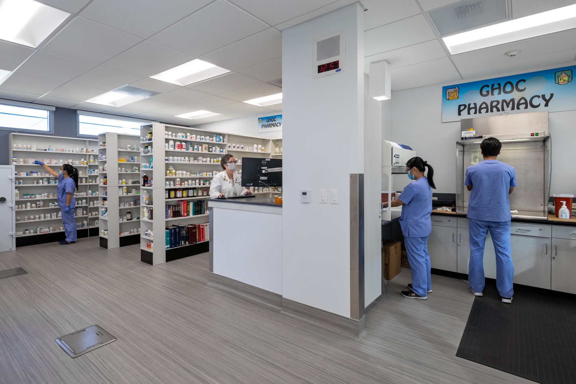 Pharmacy interior with shelves of medicines, staff in blue uniforms, and a pharmacy sign, well-lit and organized.