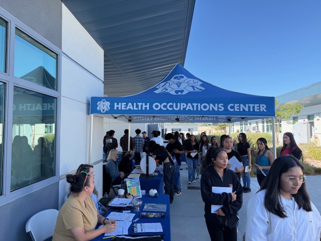 People gather outside a health occupations center under a blue tent with mountain logo, clear sky in background.