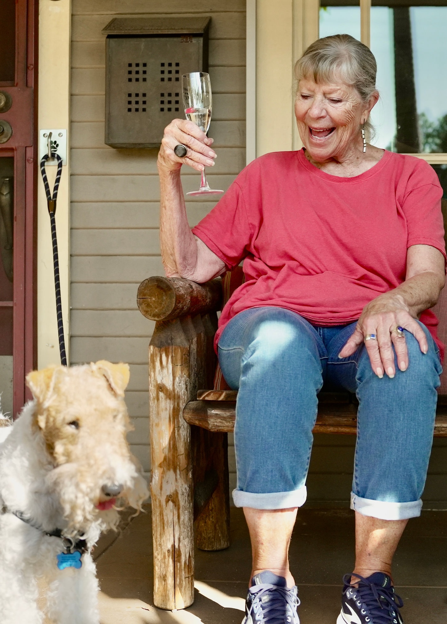 Older woman sitting on a wooden chair, holding a glass, smiling, with a dog nearby outside a house.