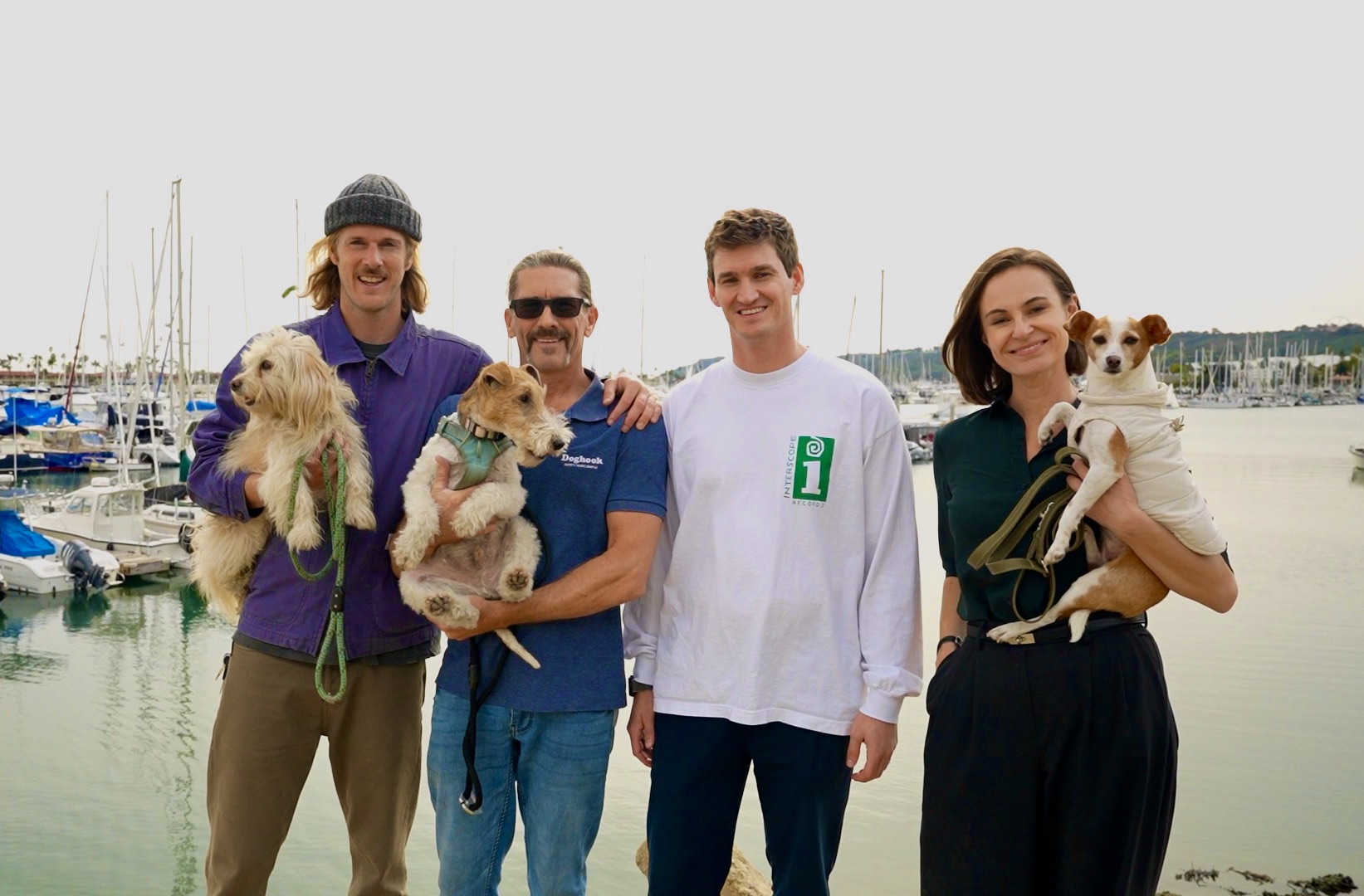 Four people stand by a marina holding dogs, with boats and water in the background, smiling at the camera.