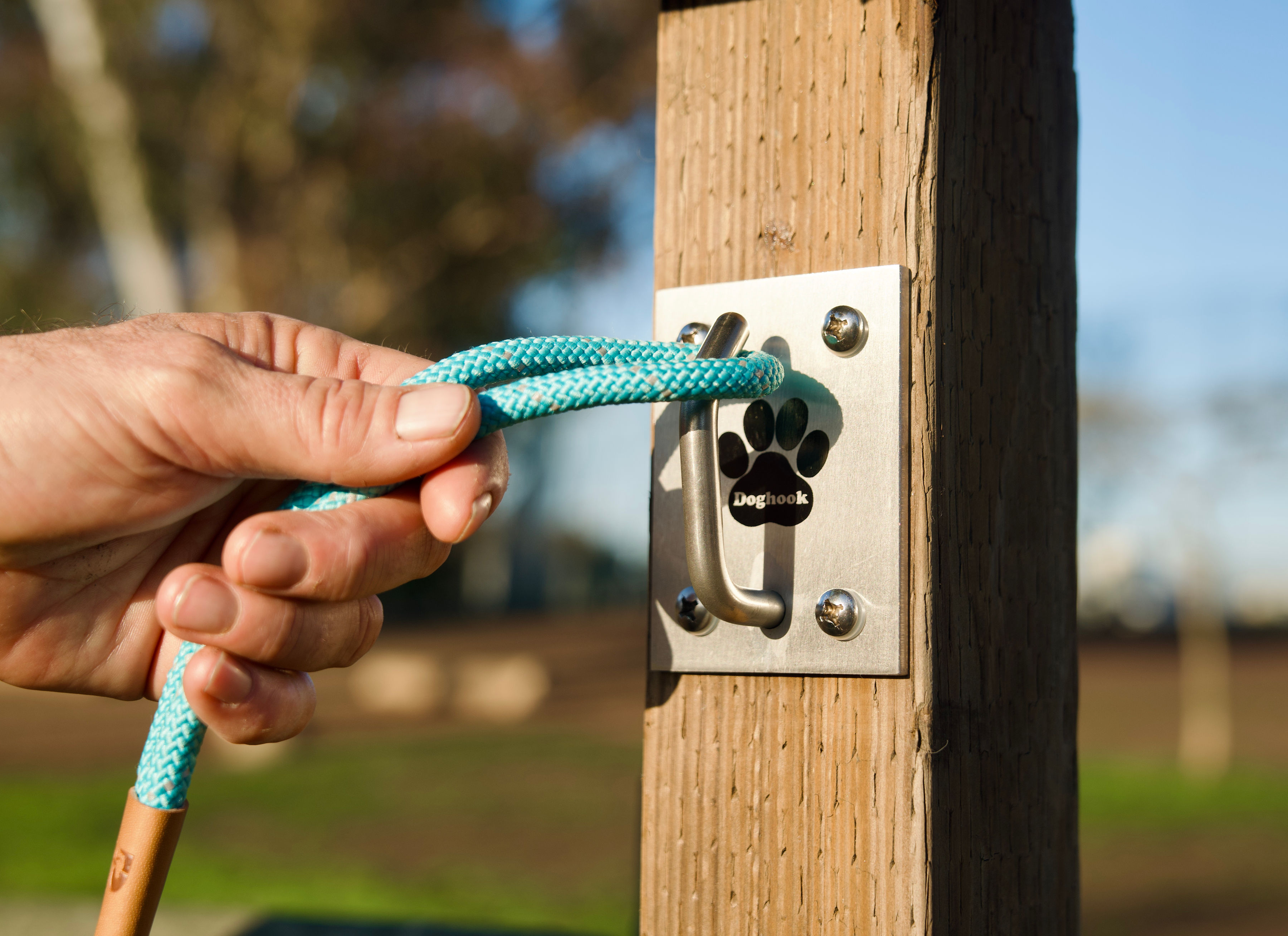 Person inserting a blue rope into a metal lock on a wooden post outdoors, with trees and sky in background.