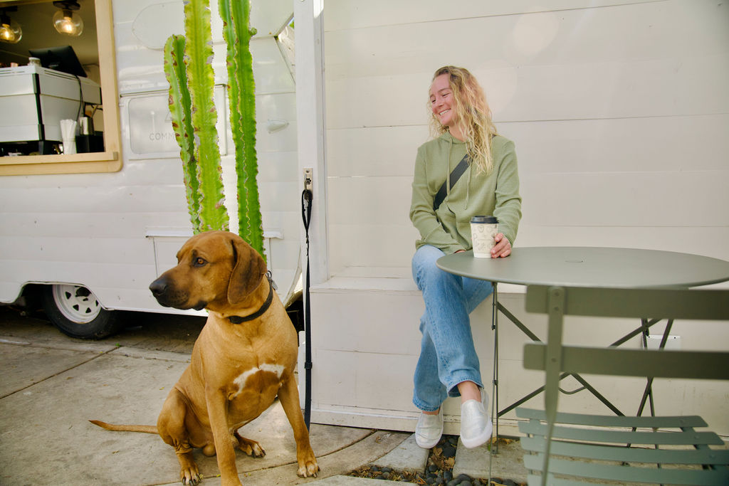 Woman sitting at a table with a mug, dog sitting nearby, white wall and cactus in background.