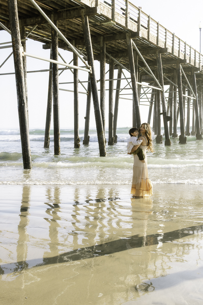 Two women stand in shallow water near a wooden pier, holding a child, with reflections on the wet sand.