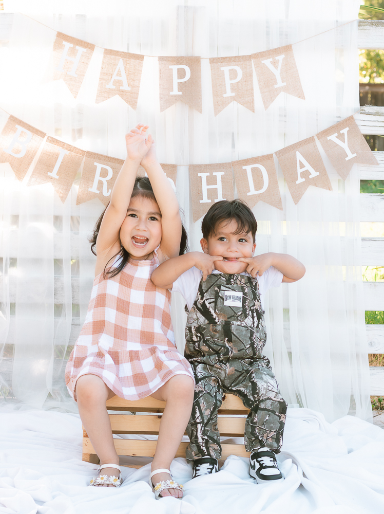 Two children sitting on small wooden crates in front of a birthday banner, smiling and celebrating, with a white curtain background.
