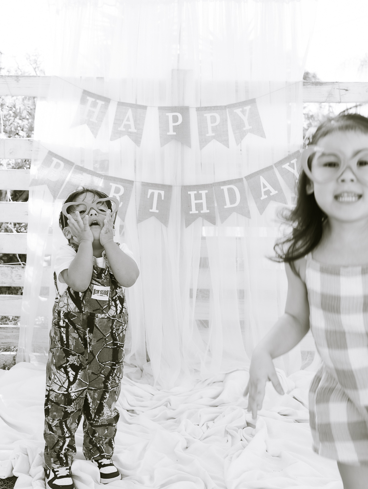 Two children celebrating birthday with a banner in the background, one smiling and the other covering their face, in a decorated room.