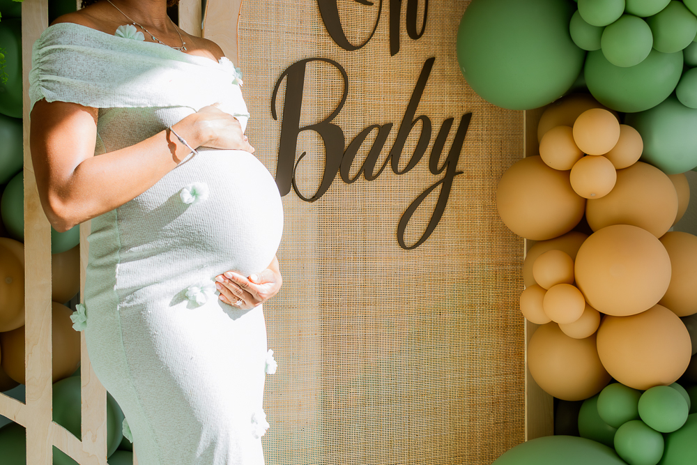 Pregnant woman in white dress holding her belly at a baby shower with balloons and a sign that says