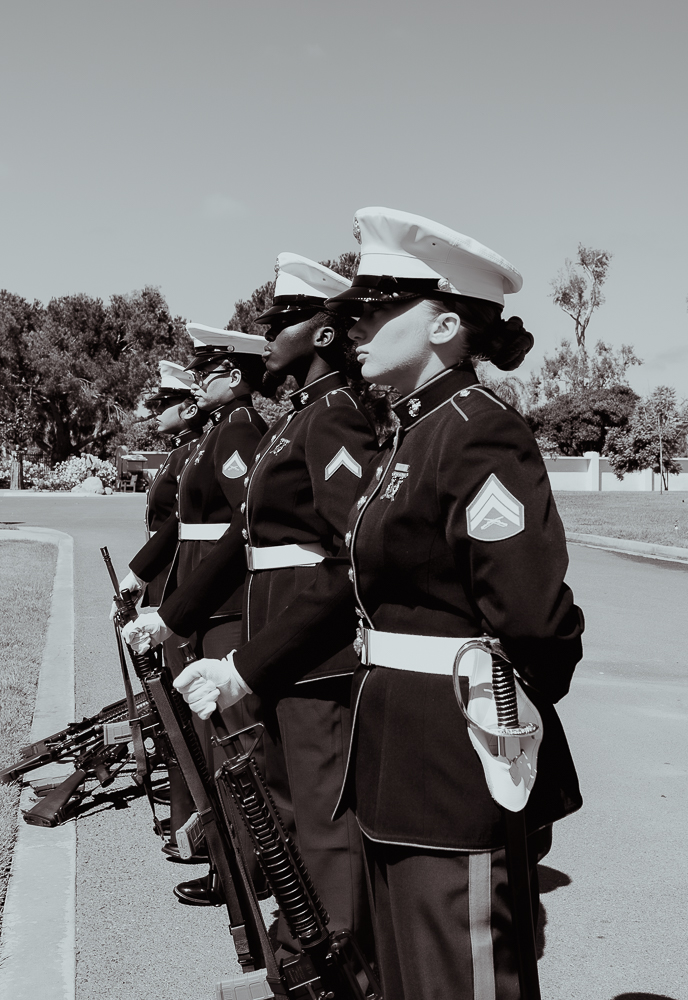 Four uniformed women in military dress standing in a line outdoors, holding rifles, with trees and a clear sky in the background.