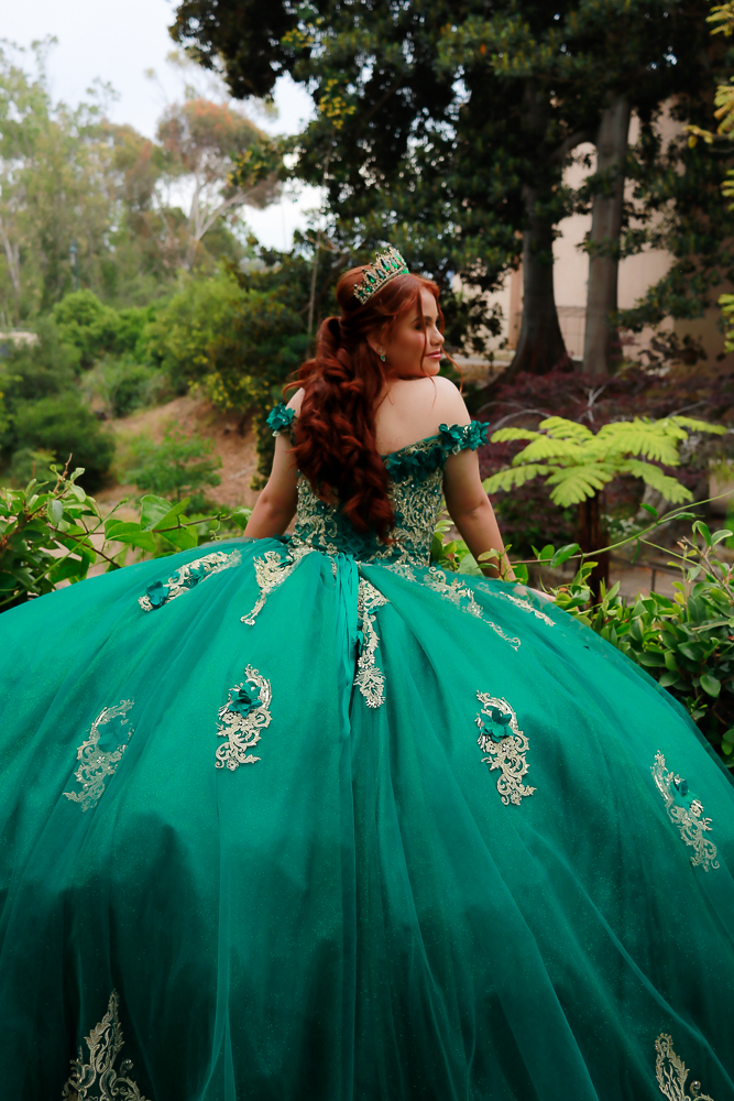 Woman in a teal gown with lace details, standing outdoors among trees and greenery, facing away from the camera.