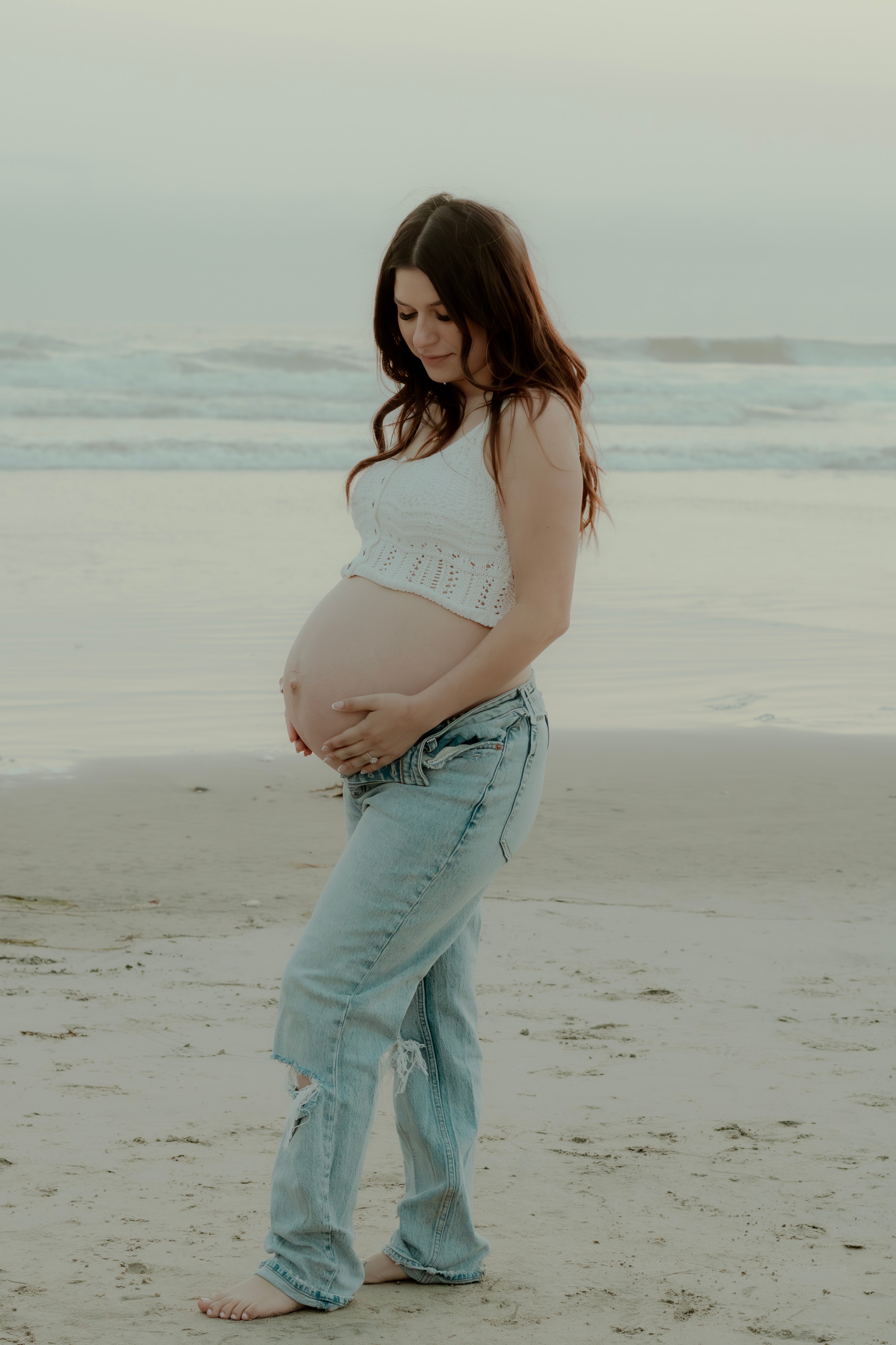 Pregnant woman standing on beach, looking down at her belly, with ocean waves in background.