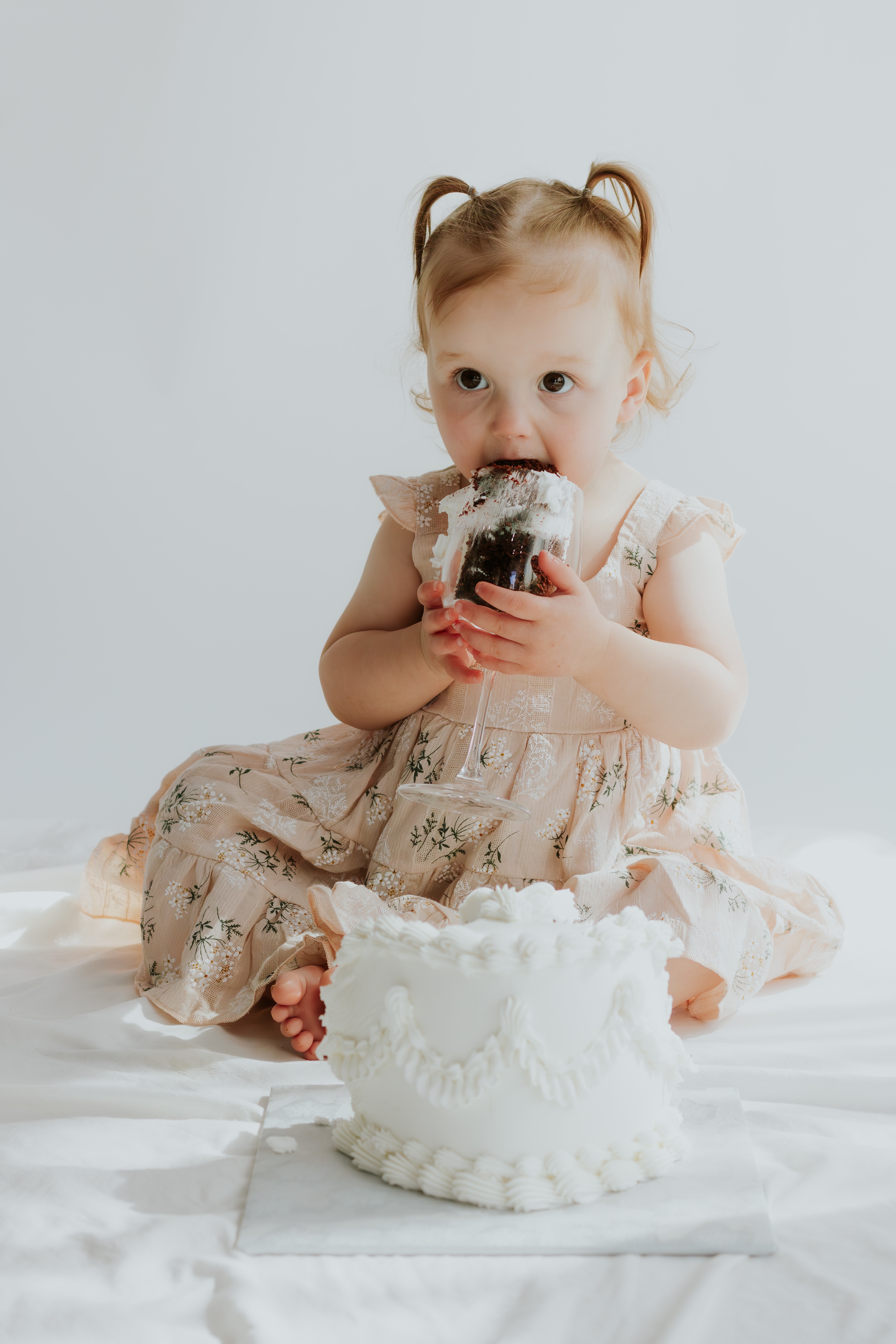 Young girl with light hair in pigtails, wearing a dress, eating cake from a bowl, sitting on a white surface.
