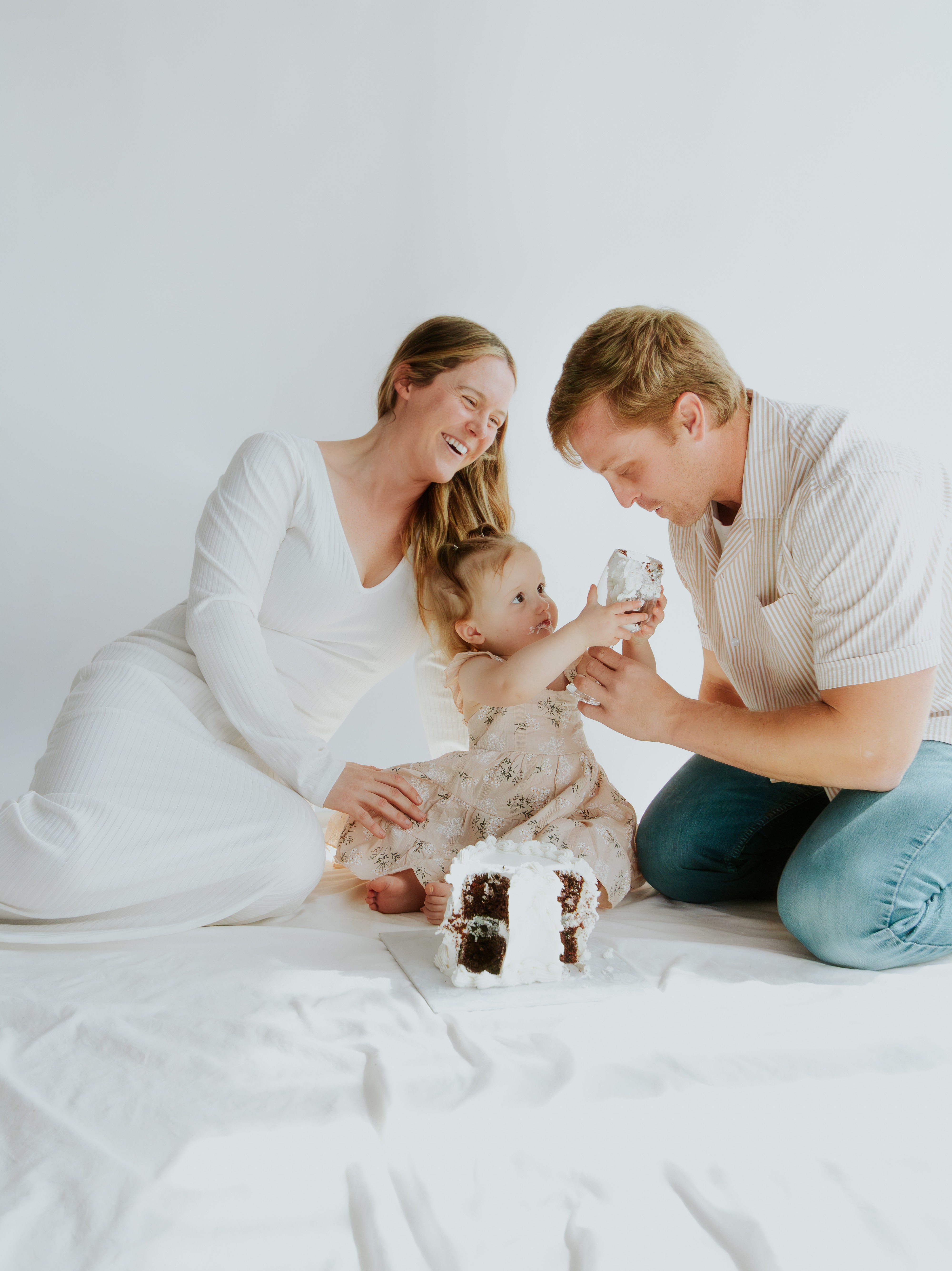 Family of three with a small dog sitting on a white sheet, smiling and playing together, against a plain white background.
