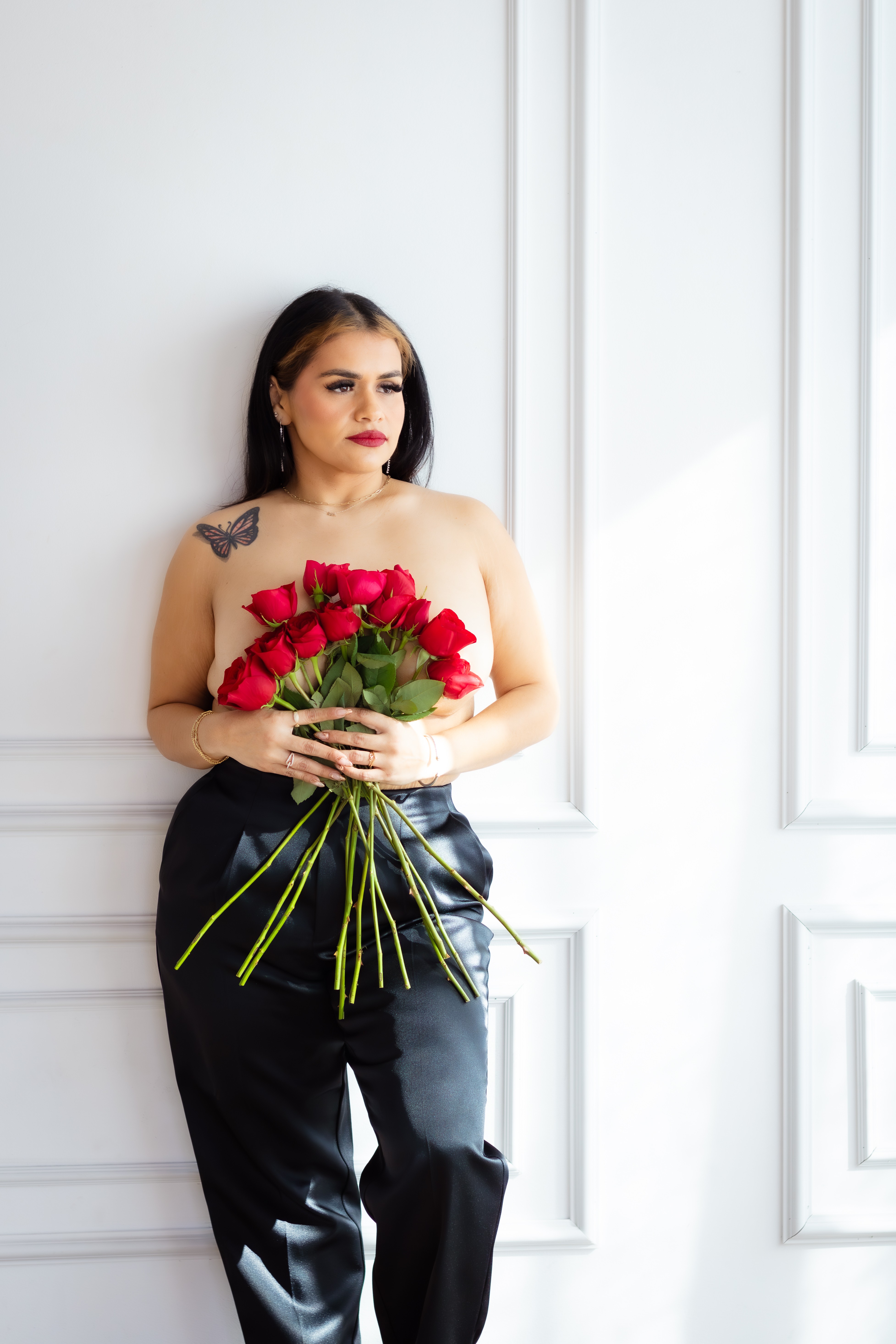 Woman with dark hair holding a bouquet of red roses, standing indoors near a window, wearing a black skirt, looking to the side.