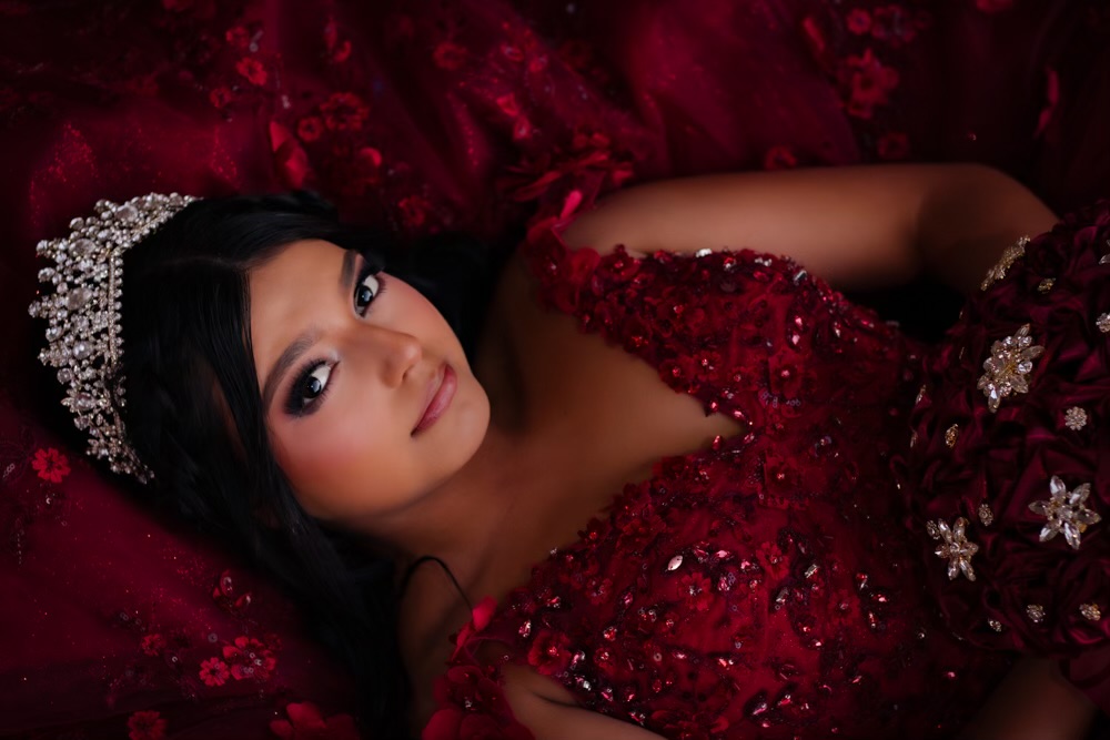 Woman lying on red fabric, wearing a red dress with embellishments and a tiara, looking at the camera.