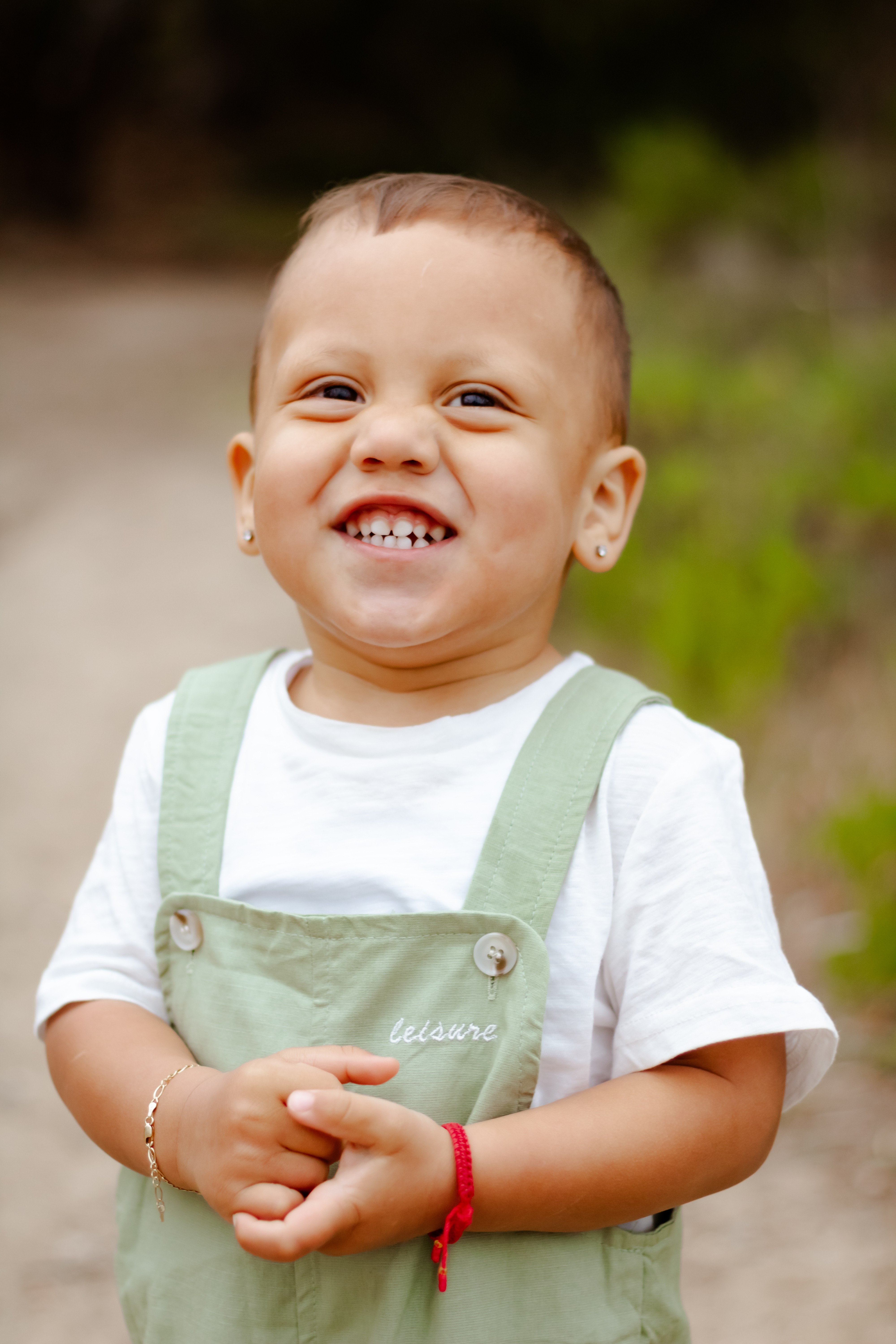 Young boy smiling outdoors, wearing a white shirt and green overalls, with hands clasped, blurred natural background.