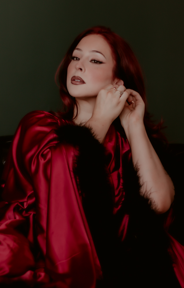 Woman with red hair wearing a red satin and black fur outfit, posing with hand near face, dark green background.
