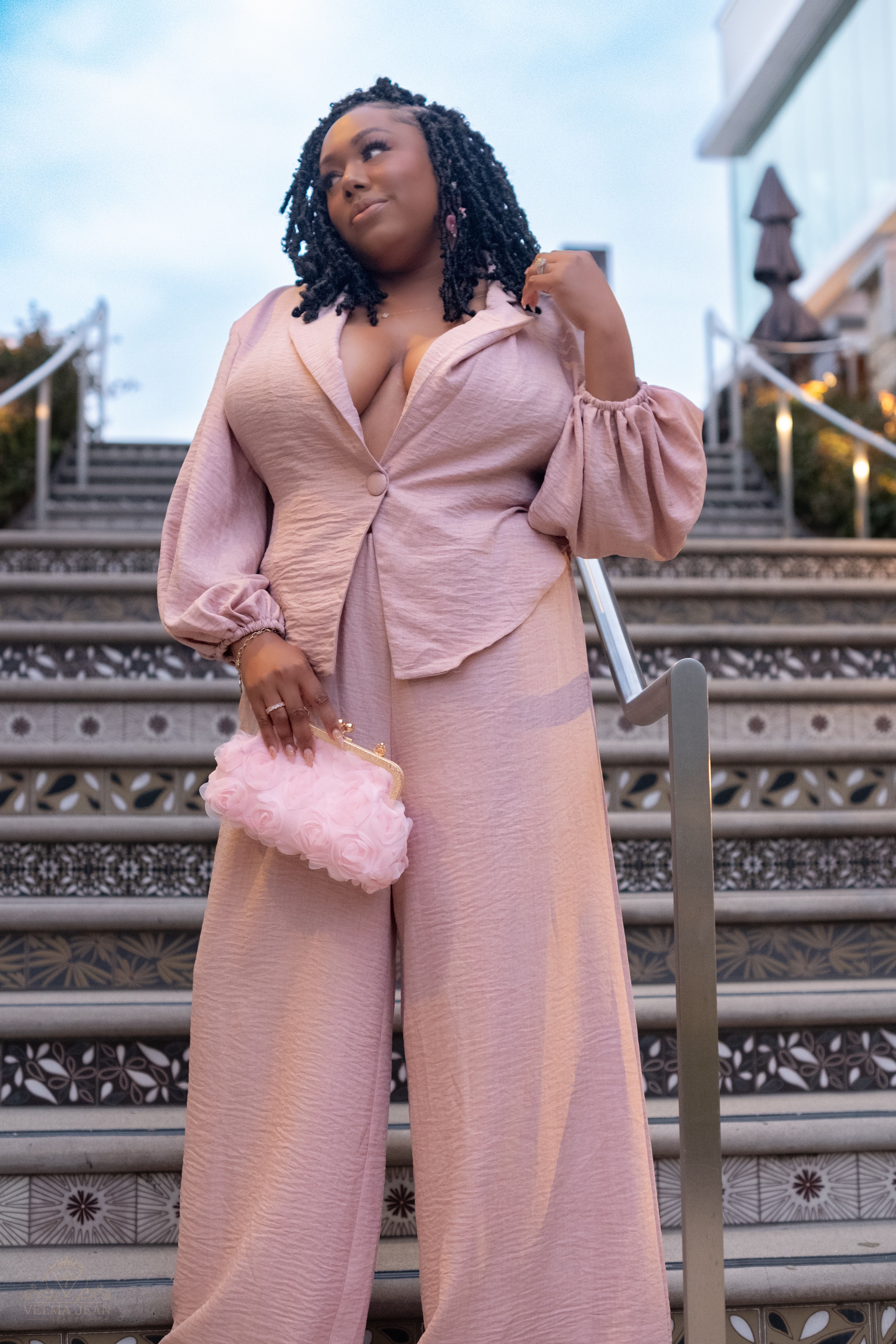 Woman in pink outfit holding a pink fluffy clutch on outdoor stairs, looking to the side.
