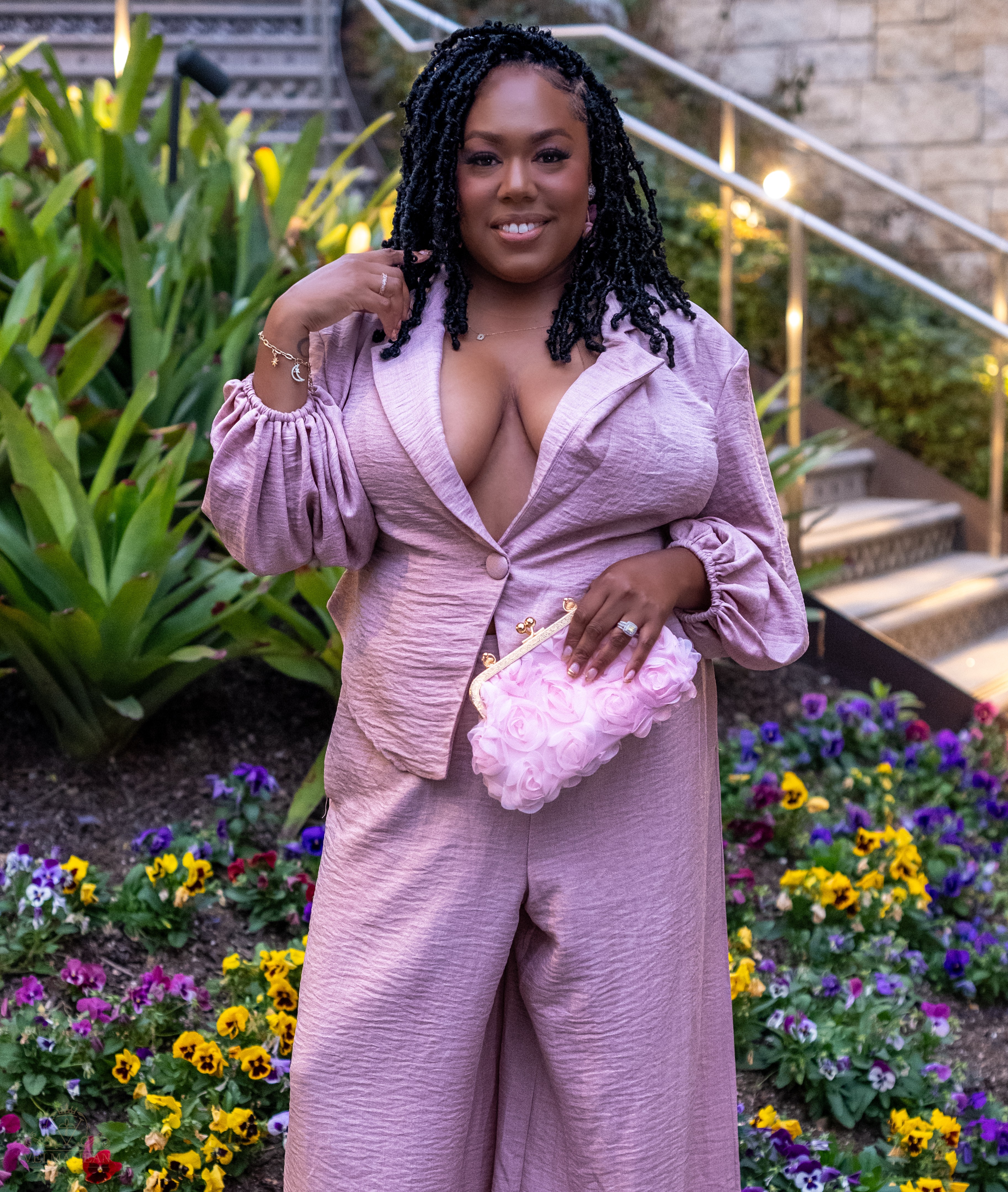 Woman in pink outfit holding a pink clutch, standing outdoors with colorful flowers and greenery.