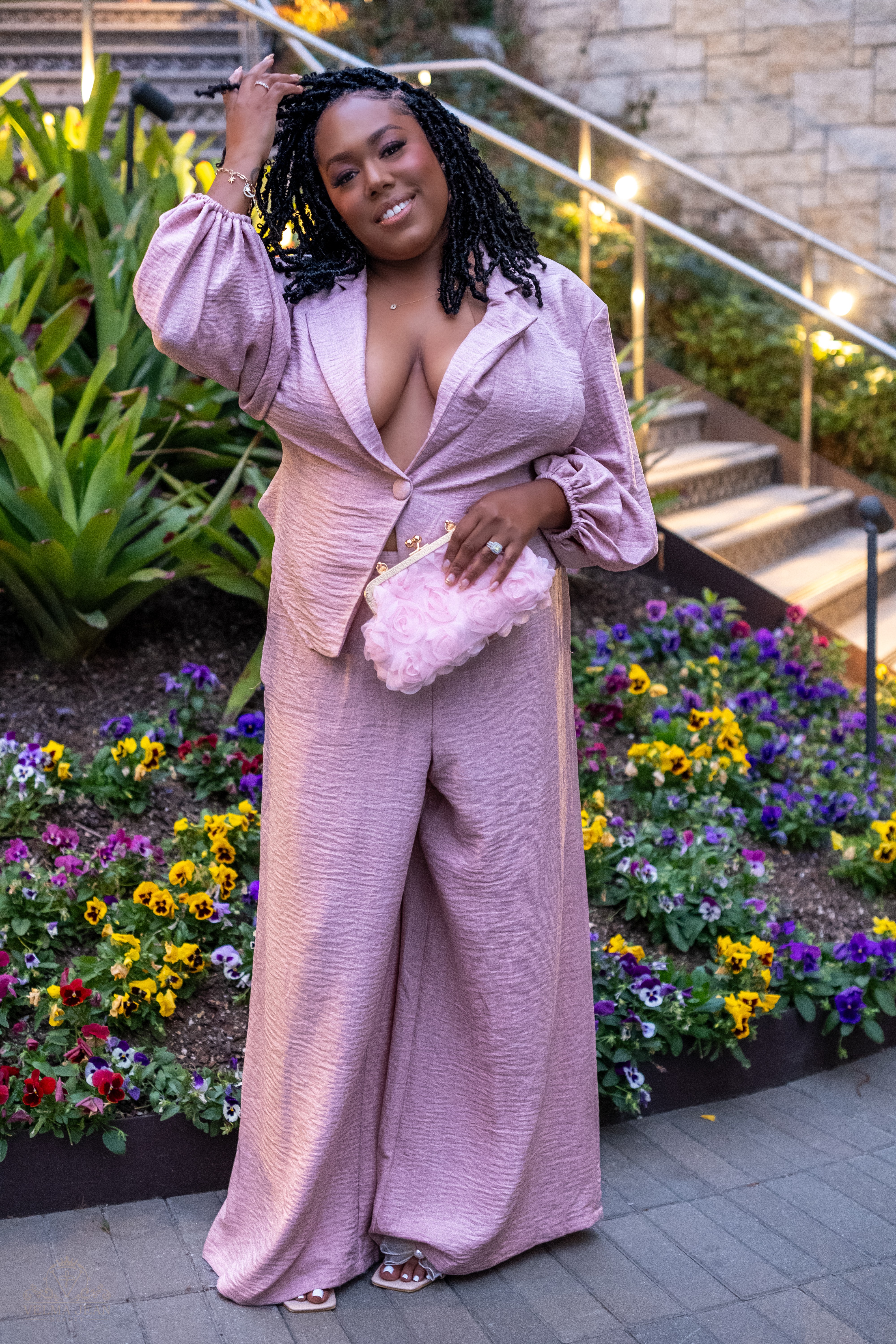 Woman in pink outfit standing outdoors near colorful flower bed and stone stairs, smiling, with hand on her head.