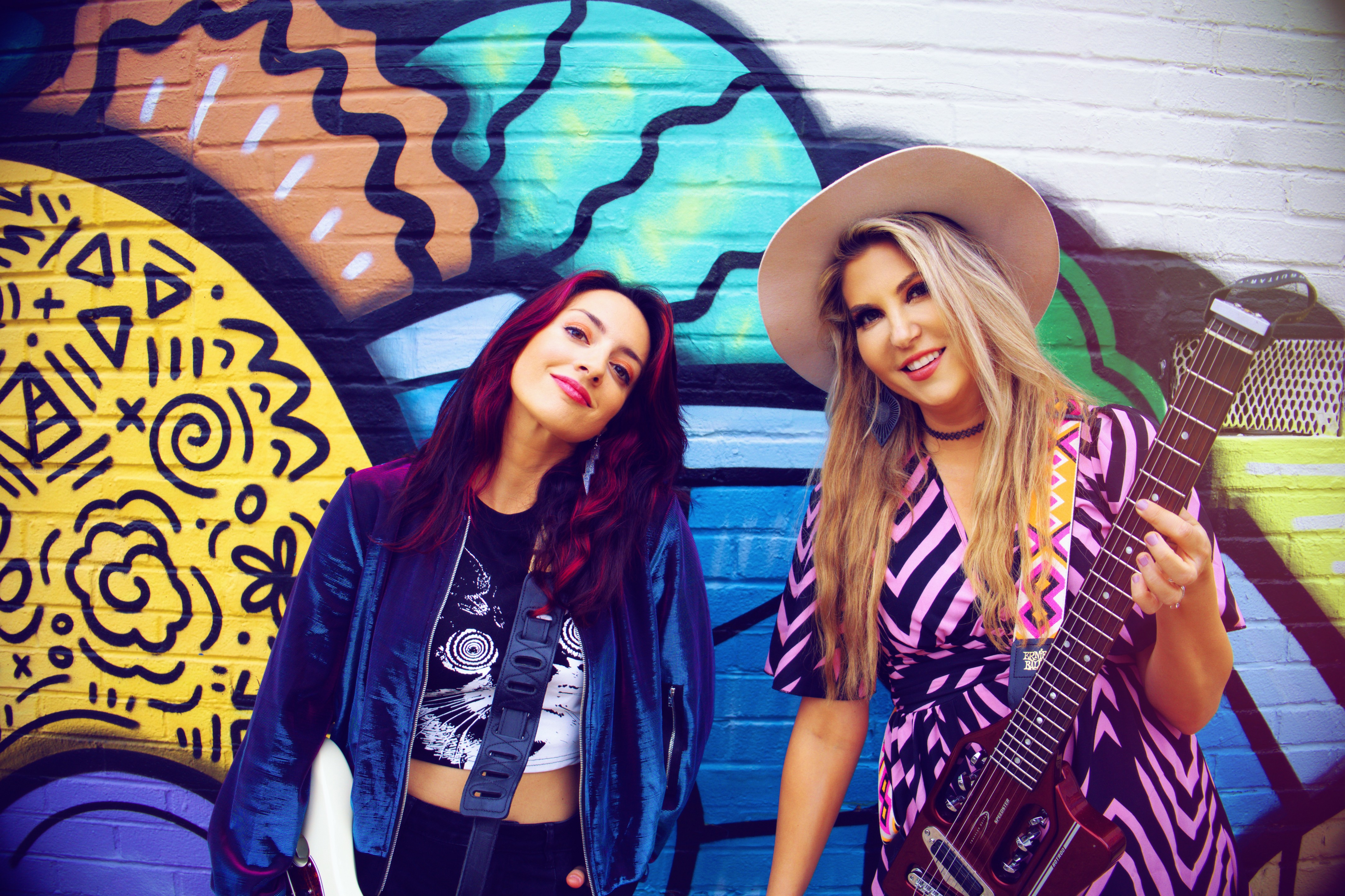 Two women stand in front of a colorful graffiti wall, smiling and posing for the camera.