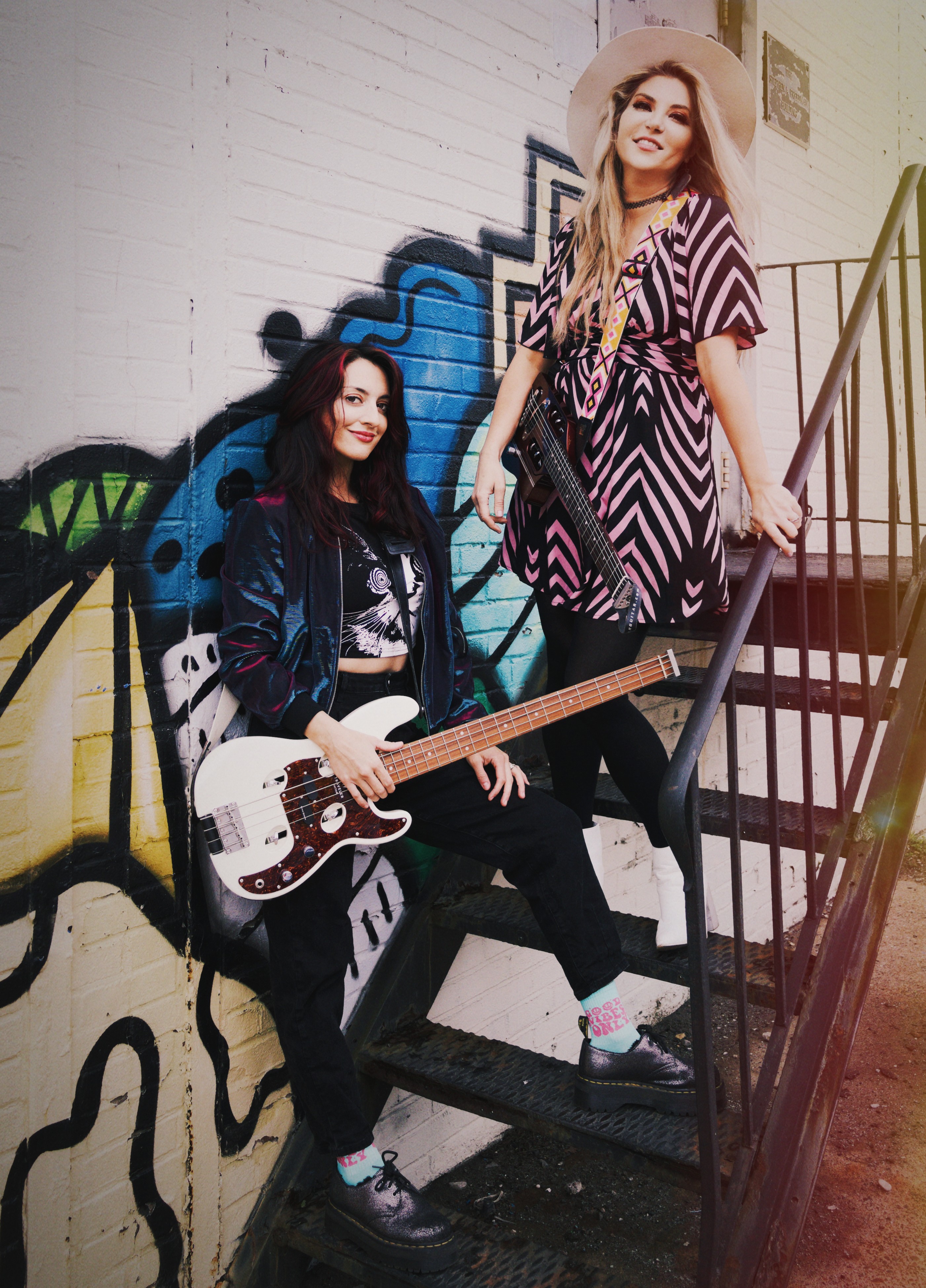 Two women with musical instruments standing on a staircase against a graffiti wall, one holding a guitar, smiling.