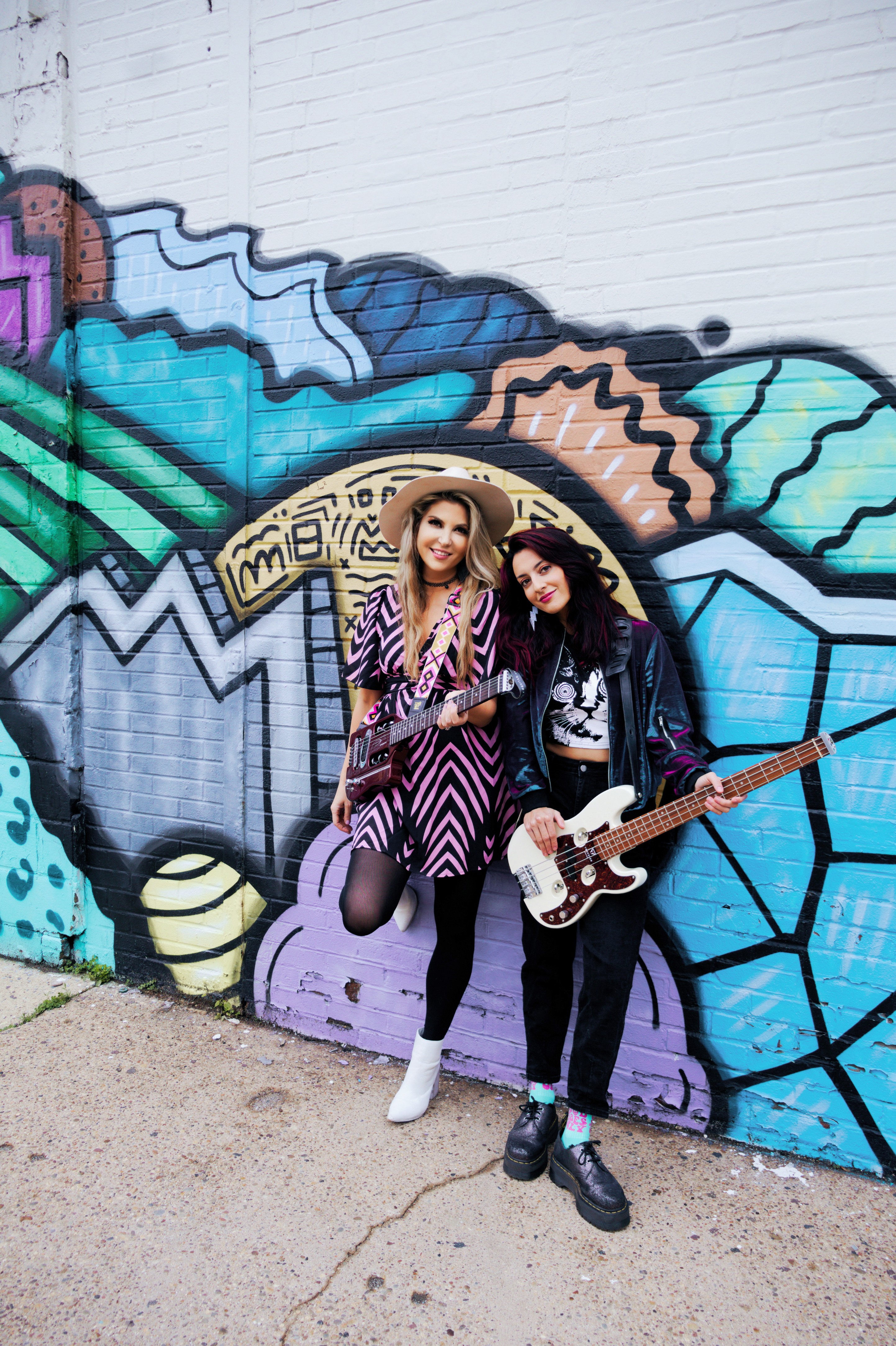 Two women stand in front of a colorful graffiti wall, one holding a tablet and the other holding a guitar.