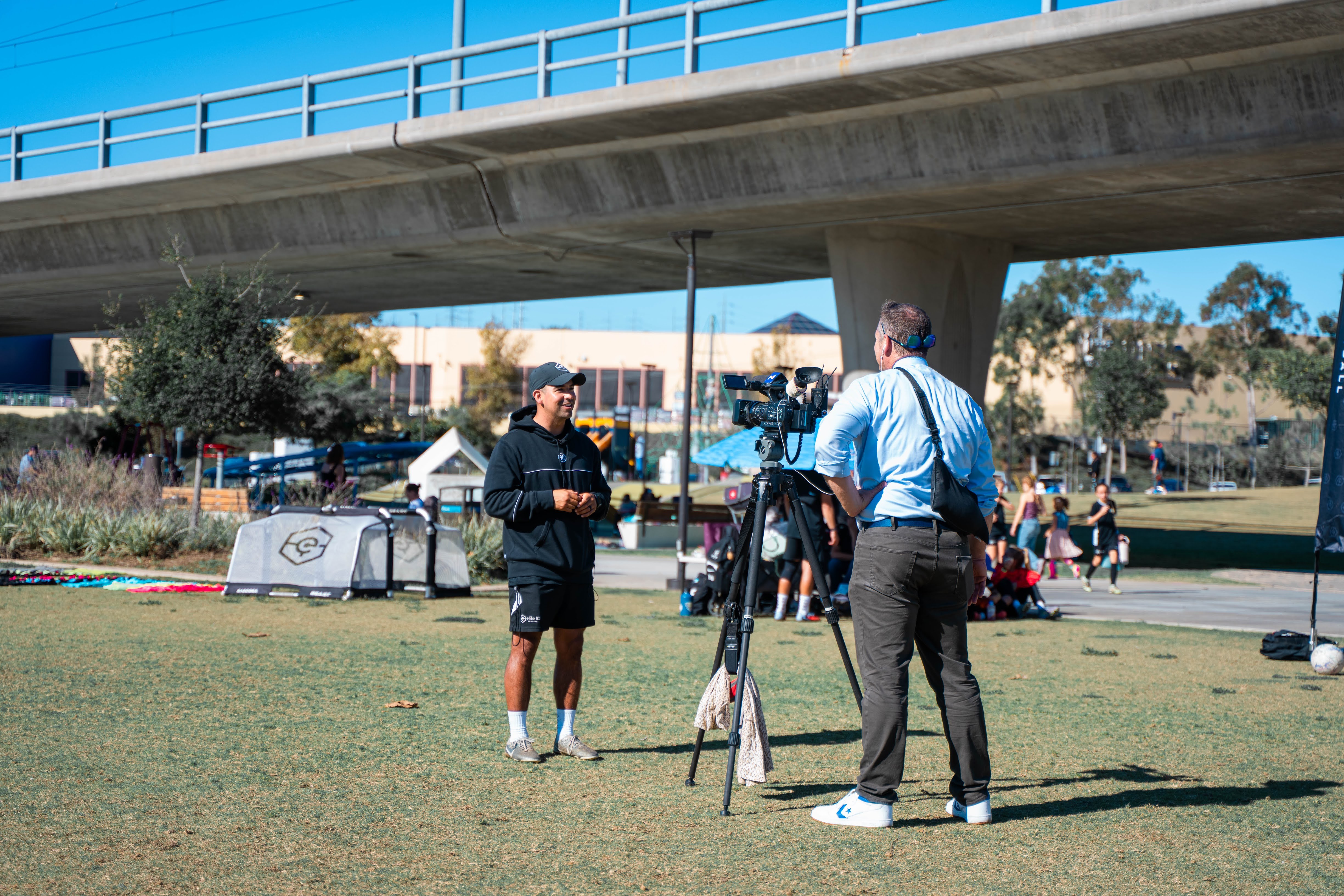 Two men are standing on grass near a camera under a bridge, with people and equipment in the background.