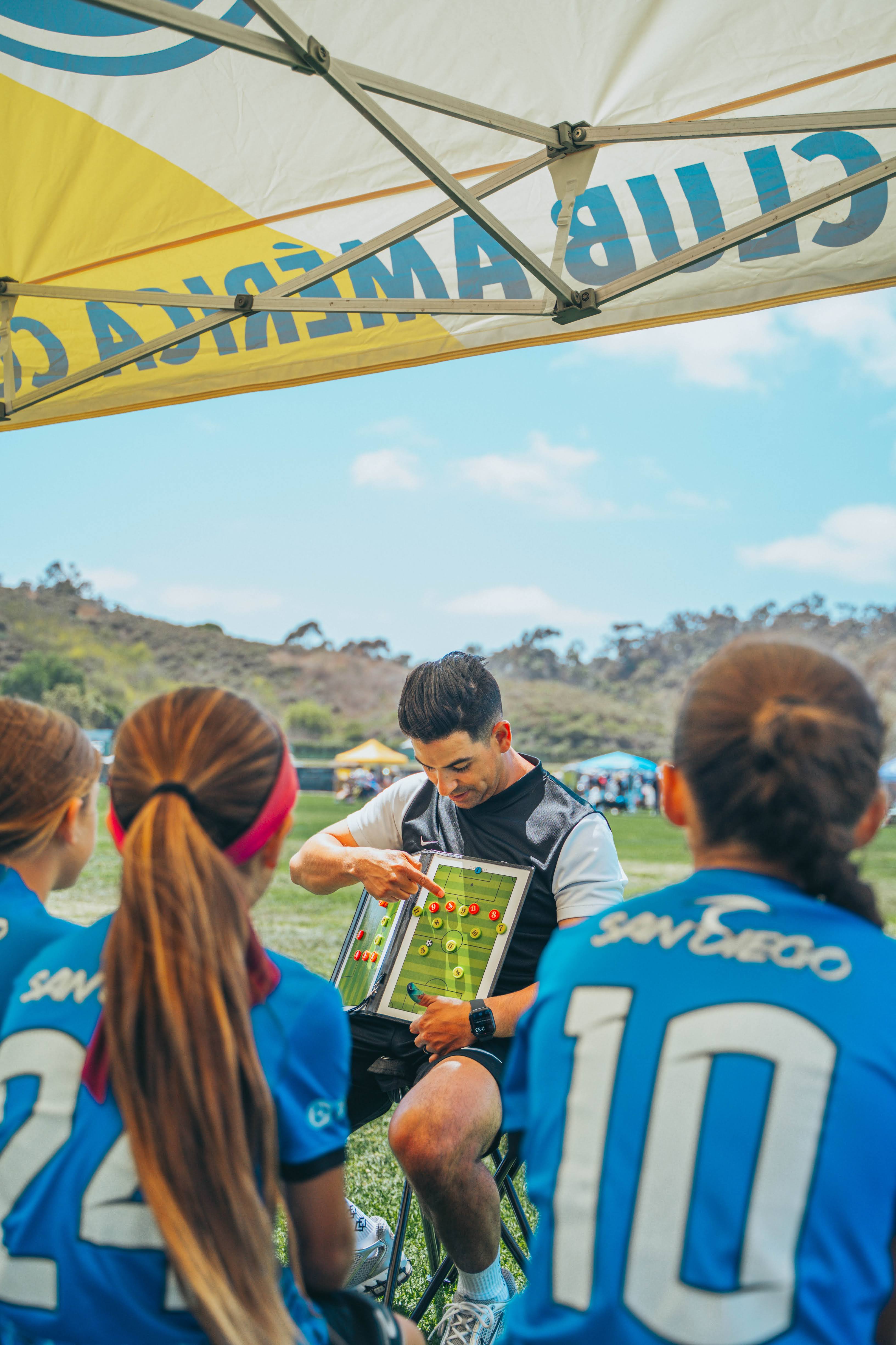 Group of young people in blue sports jerseys sitting outdoors, with a man showing a tablet, under a canopy, with trees and sky in background.