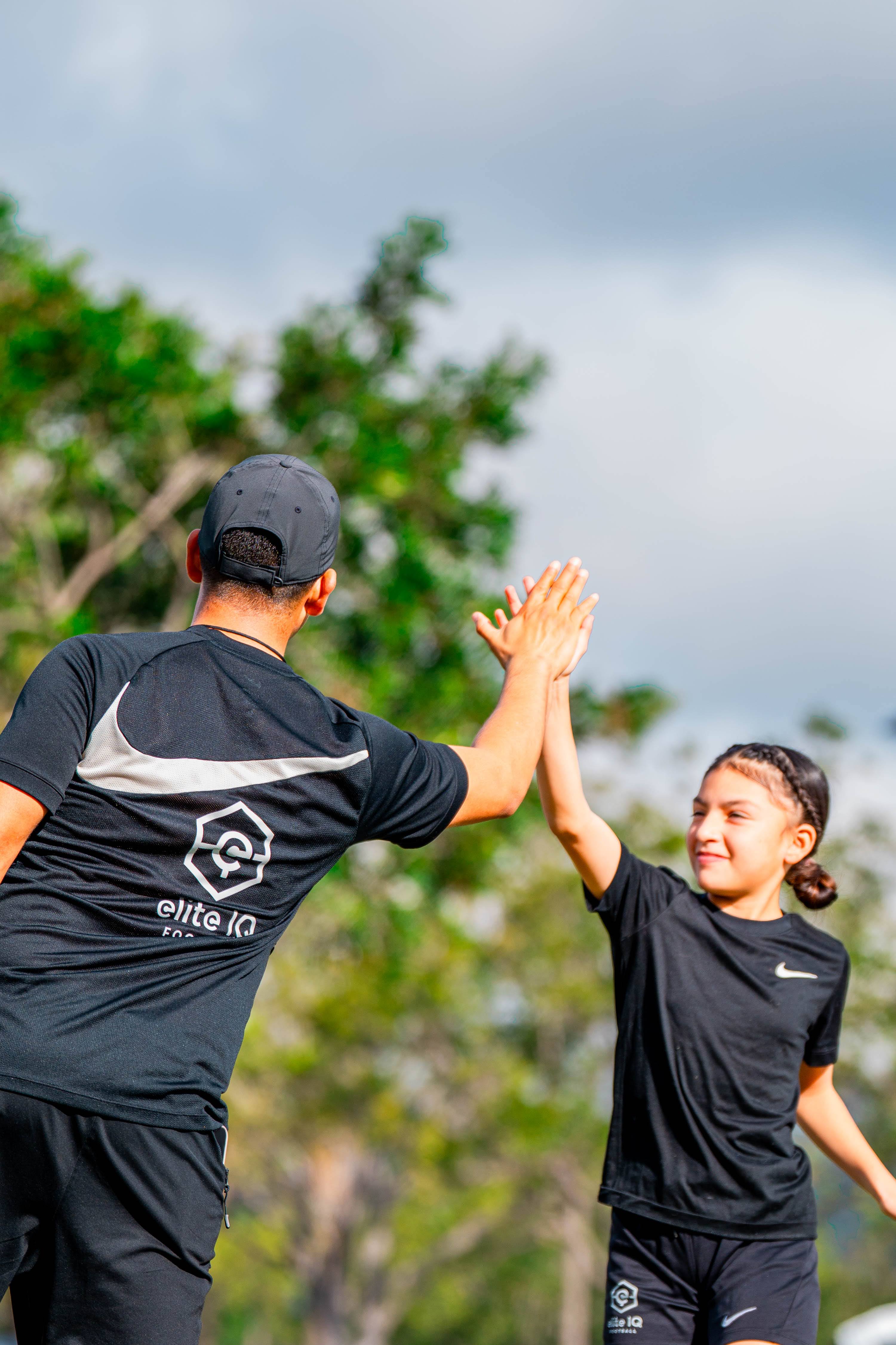 Two children high-fiving outdoors with trees and cloudy sky in background.