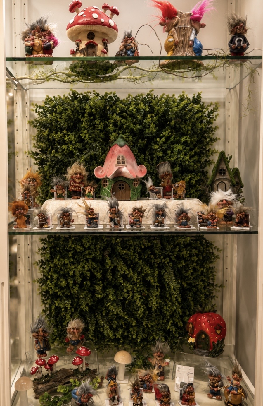 Decorative display with plants, small figurines, and colorful ornaments on glass shelves, with a pink lamp and a red ladybug figure.