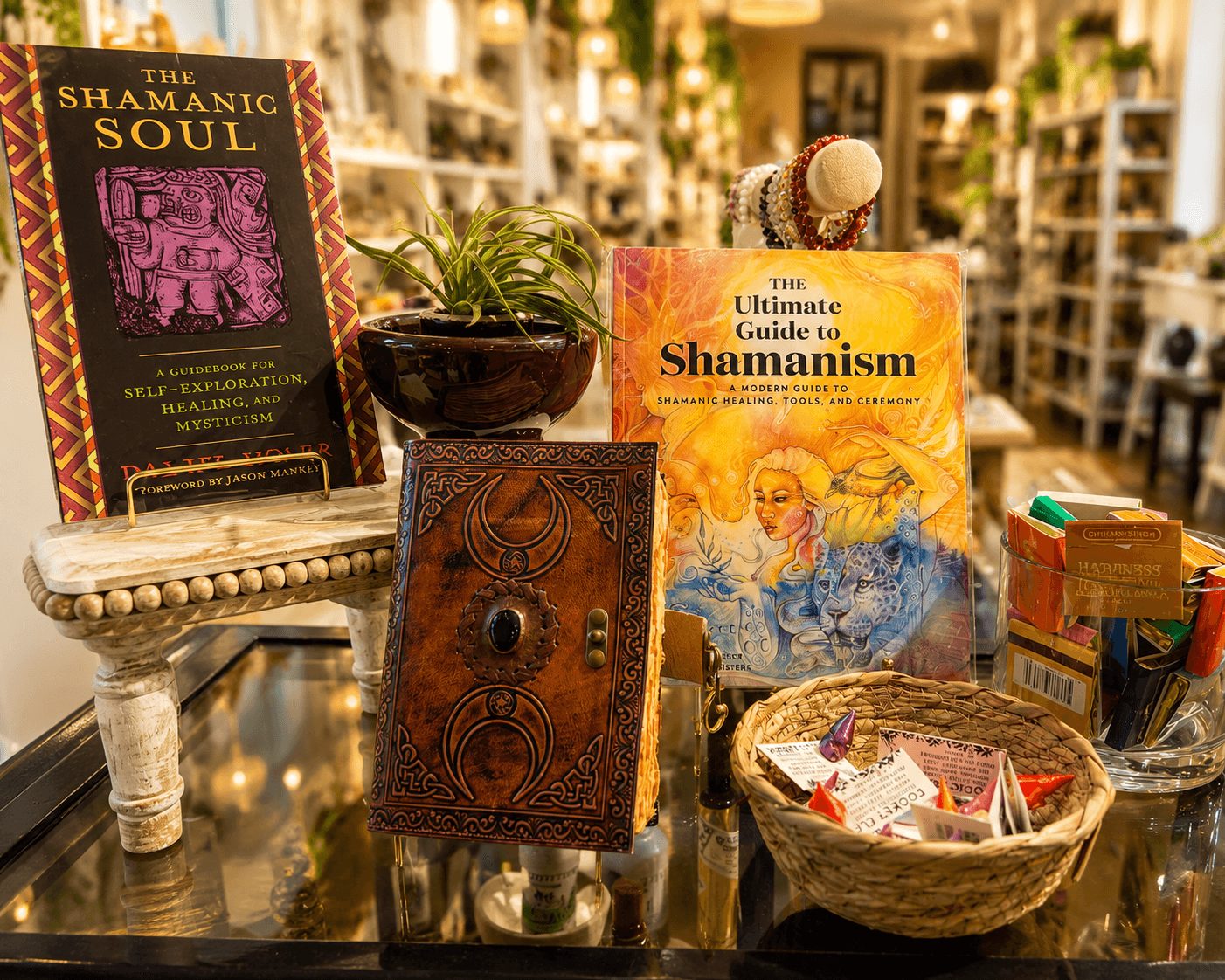 Books about spirituality and shamanism on a table with a plant, basket, and decorative items in a store setting.