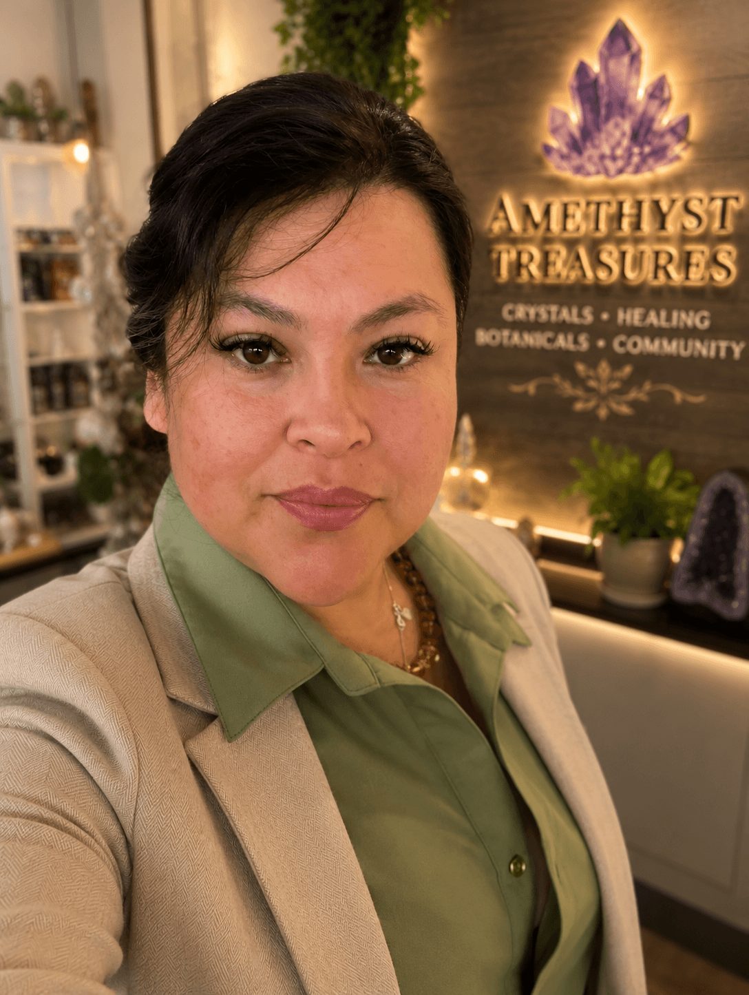 Woman with dark hair in a beige blazer and green shirt, standing indoors near a sign that reads 'Amethyst Treasures' with crystals and plants.