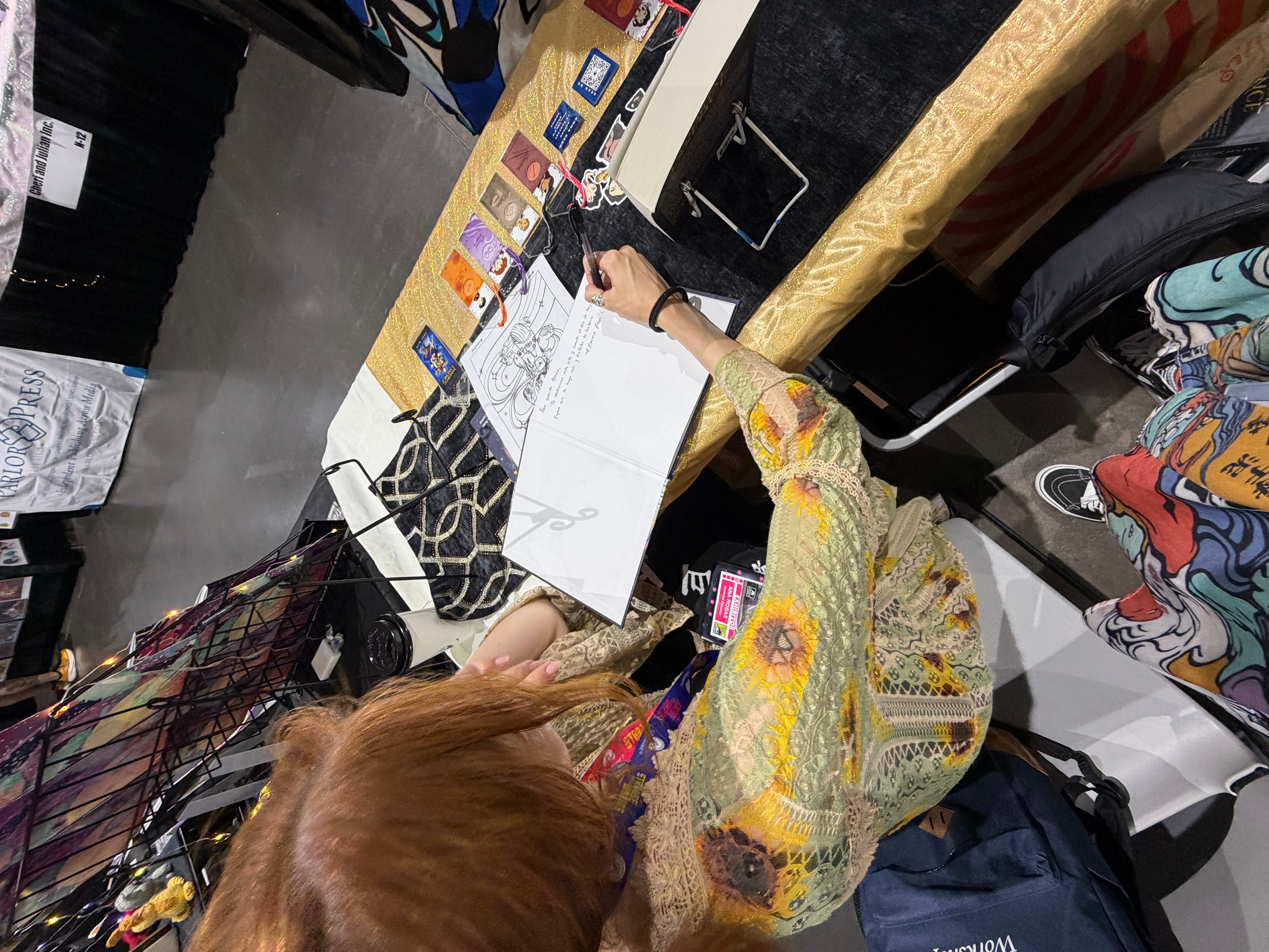 Person writing at a table with papers, bags, and various items, viewed from above.