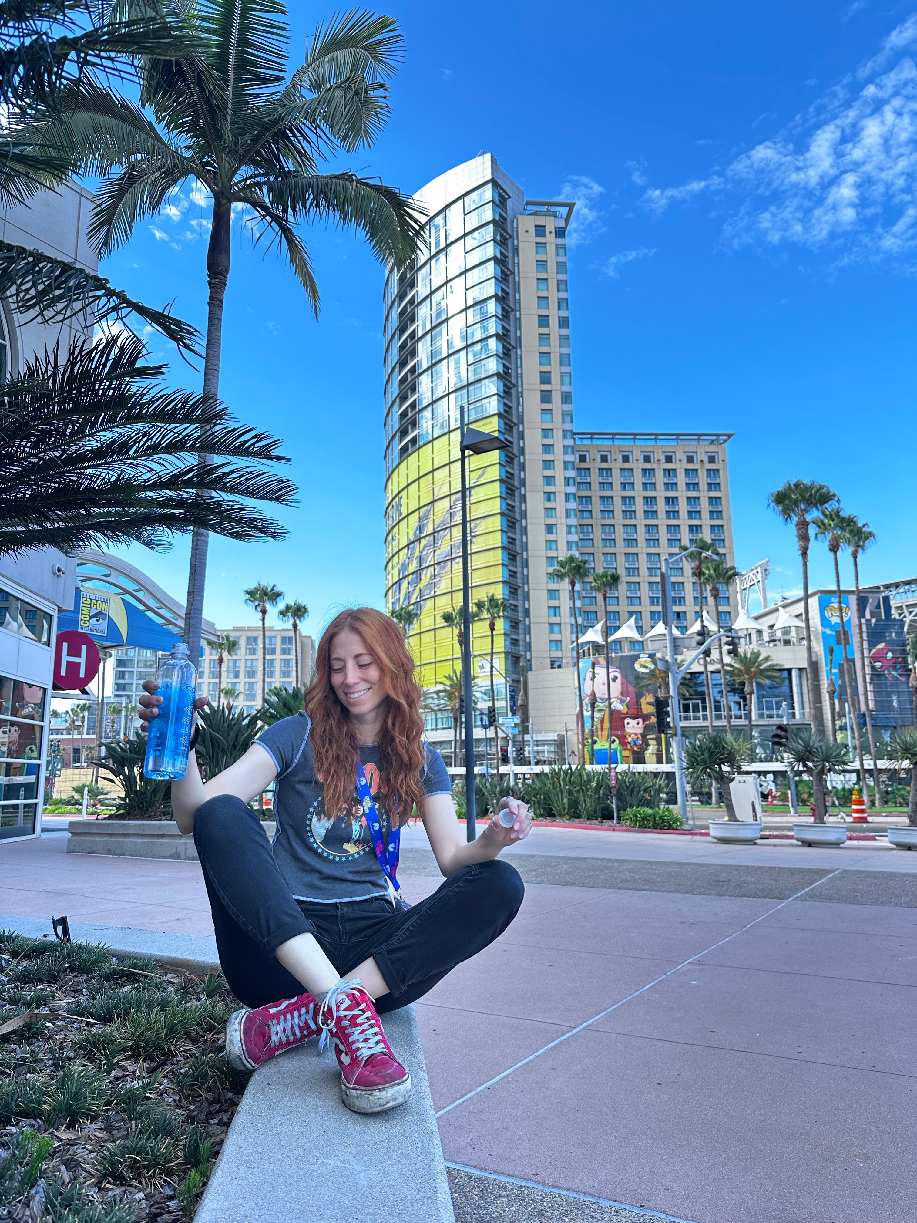 Young woman sitting on a ledge outdoors, holding a water bottle, with tall buildings and palm trees in the background.