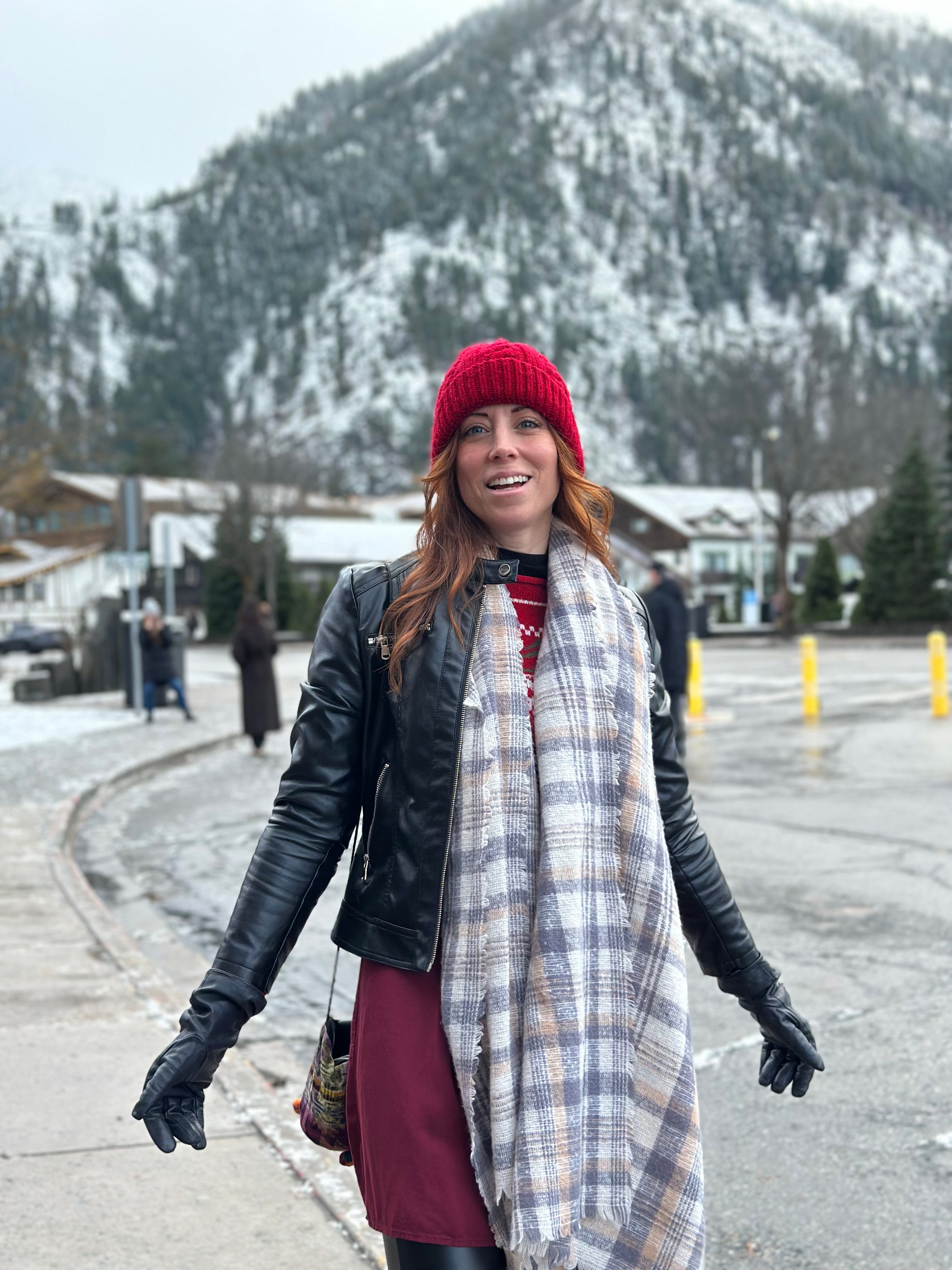 Woman in winter clothing standing outdoors with snow-covered mountain in background, smiling, wearing a red hat and black jacket.
