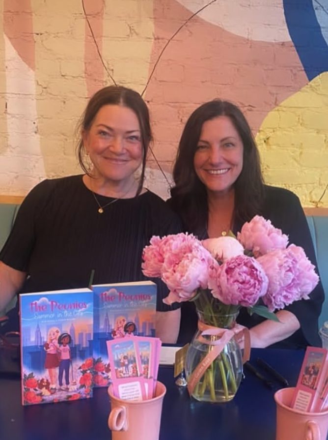 Two women smiling behind a bouquet of pink peonies, with books and pink cups in front, against a colorful mural background.