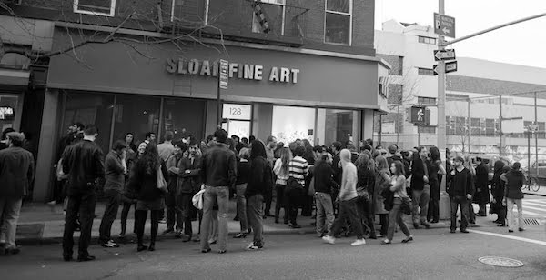 People line up outside a fine art store, some waiting and others walking by, in black and white.
