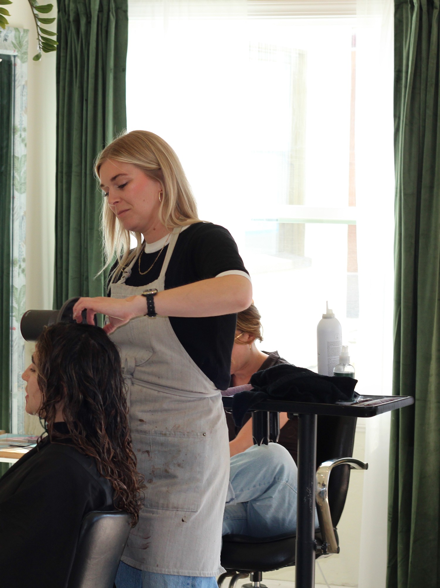 A woman with blonde hair styles a person with dark, curly hair in a salon with green curtains and natural light.