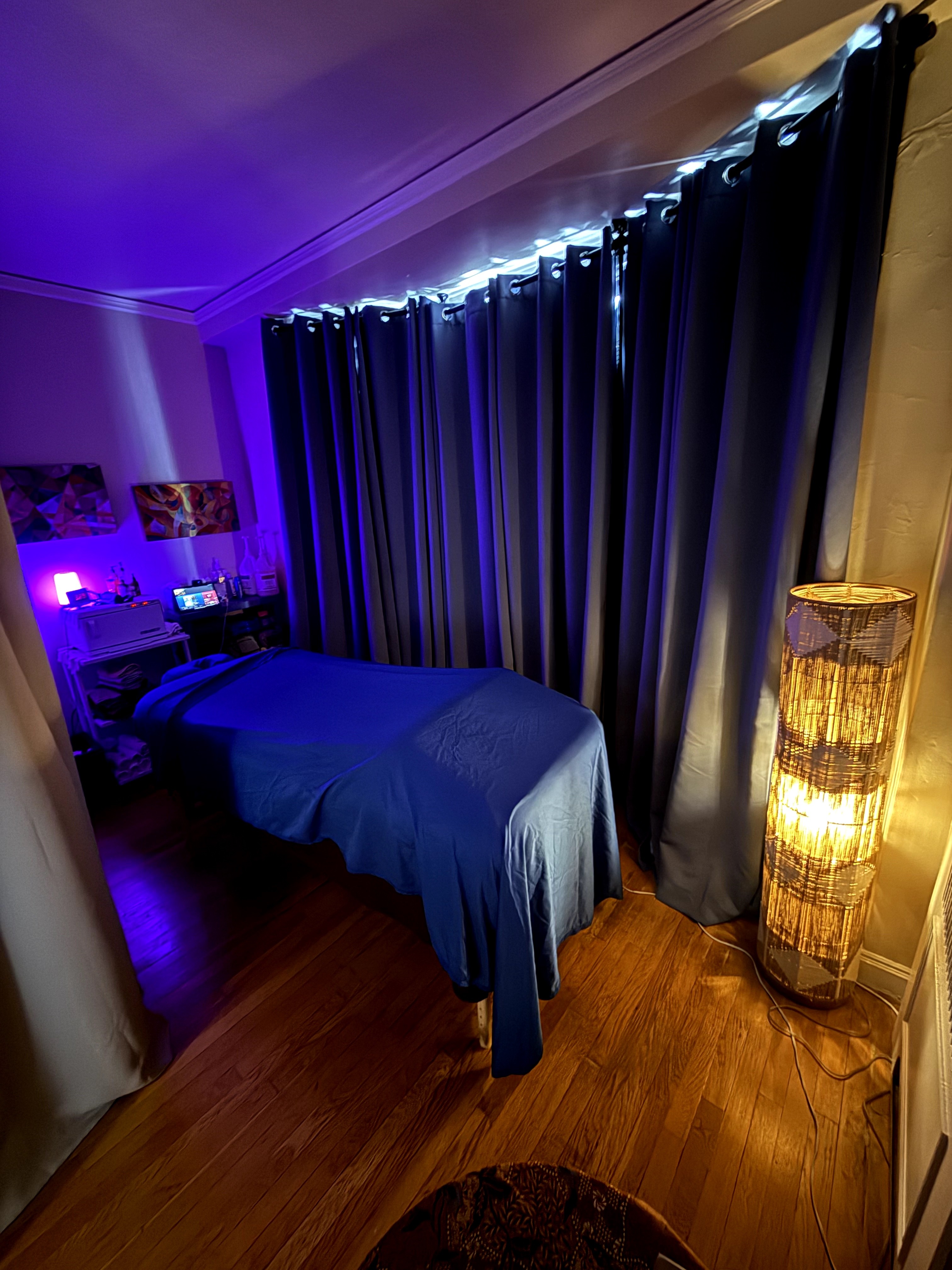 Bedroom with bed, dark curtains, and a tall lamp, illuminated by soft lighting, wooden floor, and purple accent lighting in the background.
