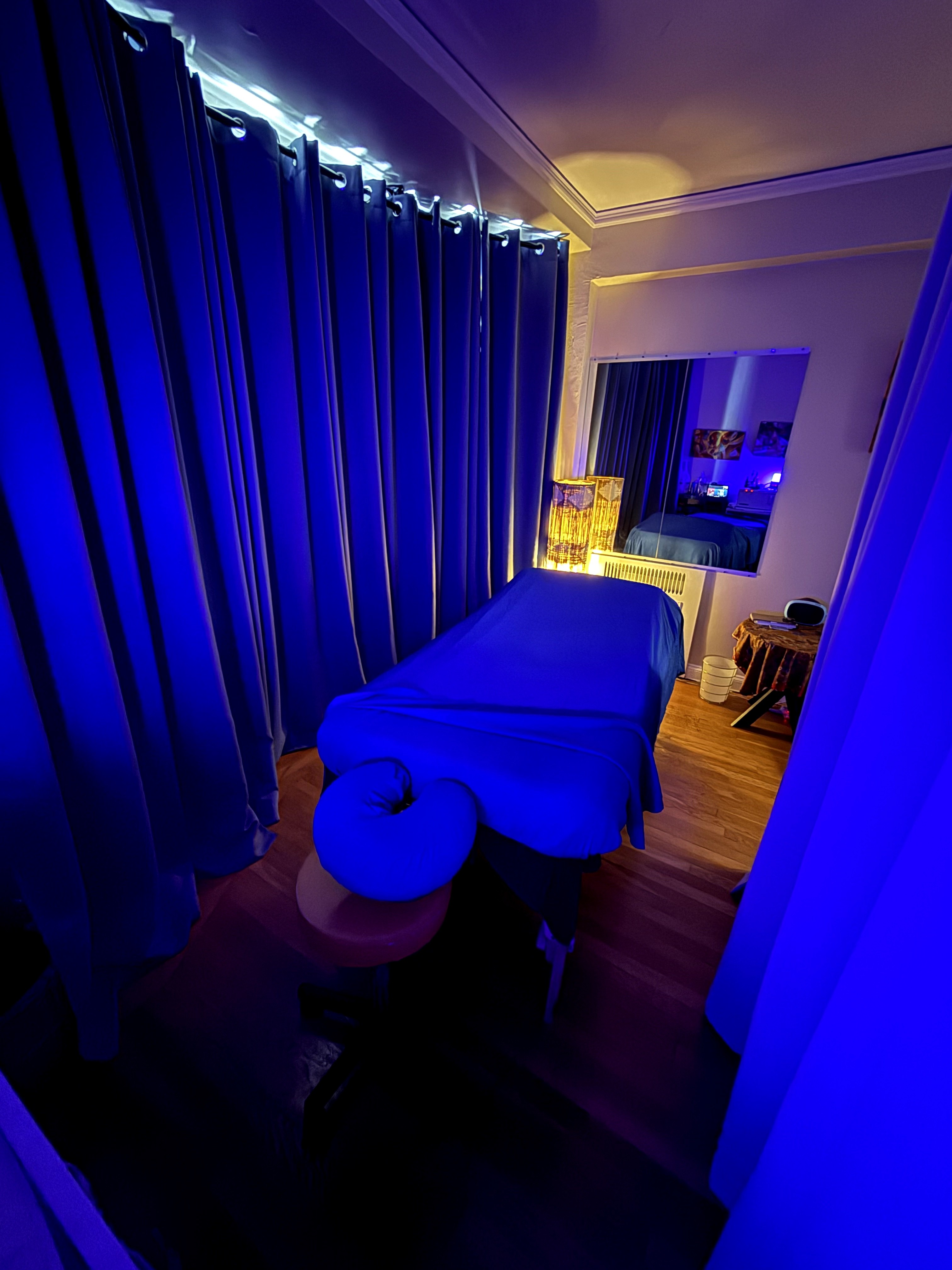 Massage table in a dimly lit room with blue curtains and a window, illuminated by blue and yellow lighting.