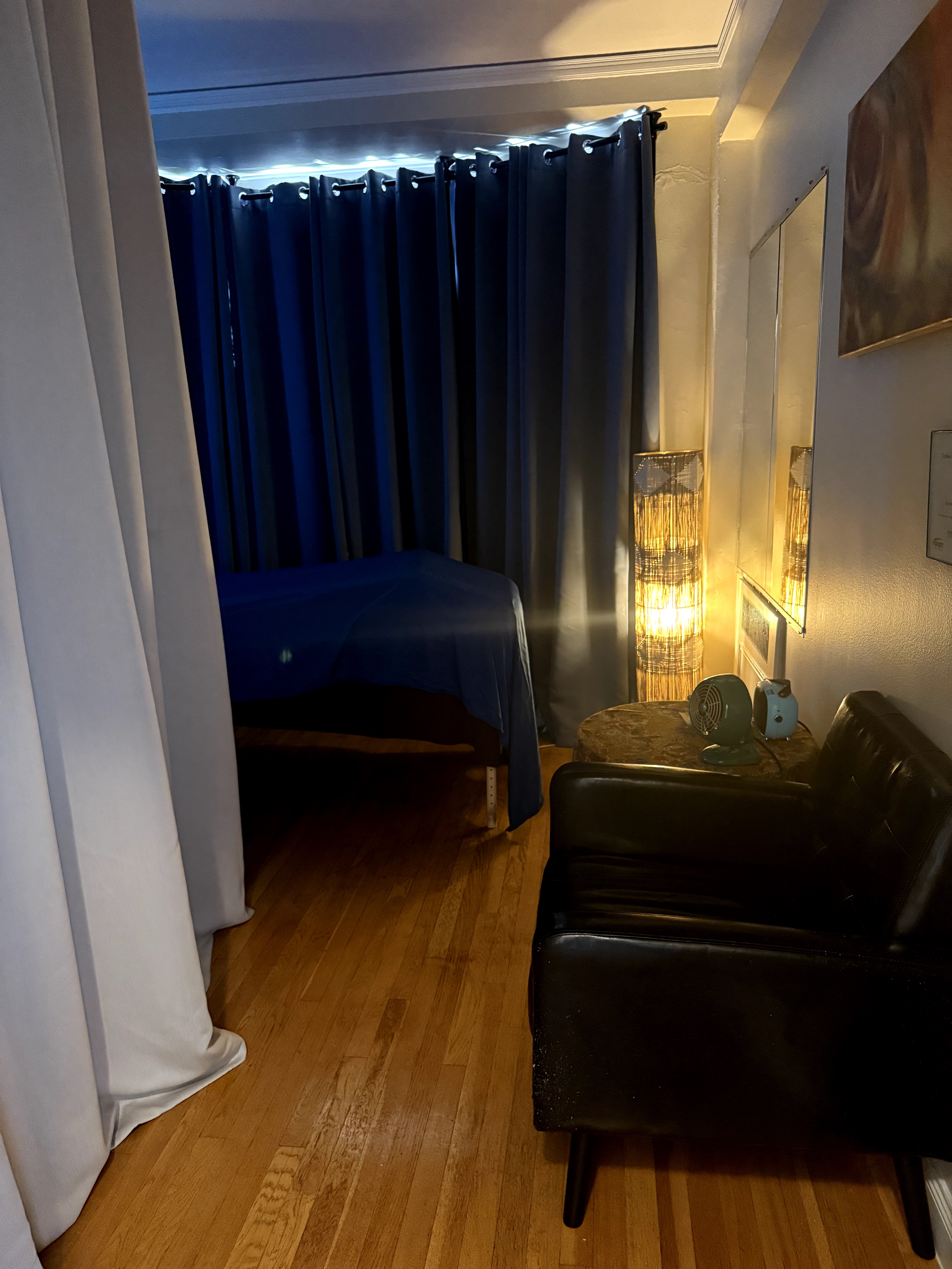 Upright bedroom with dark curtains, bed, black armchair, and a lamp on a side table, wooden floor, and wall art.