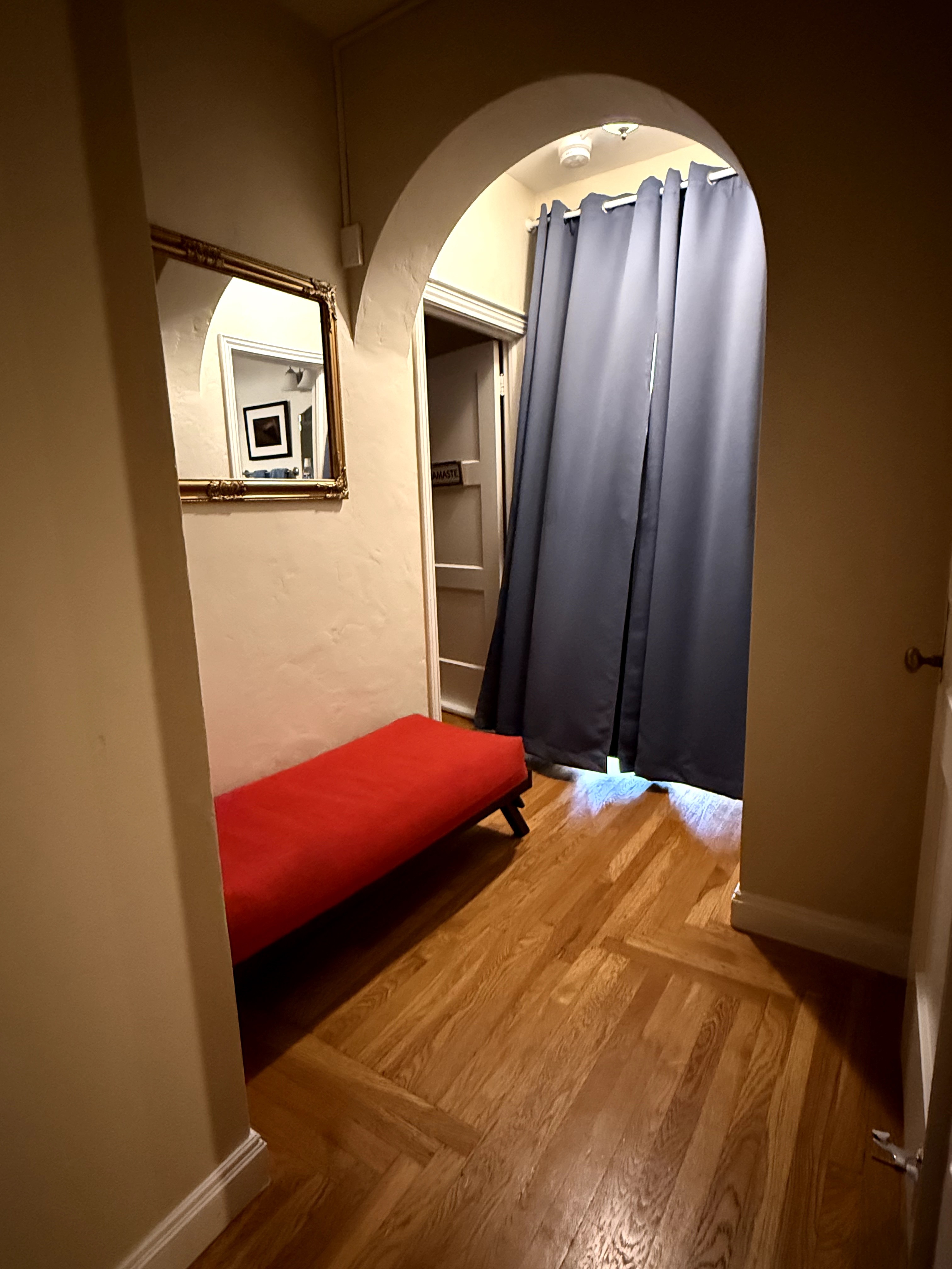 Small hallway with a red bench, mirror, and doorway with dark curtains, hardwood floor, and beige walls.